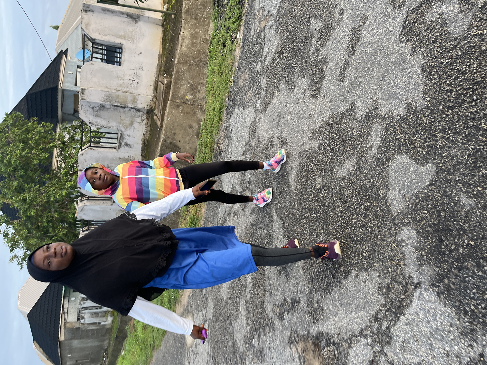 The image depicts two individuals walking on a paved road, possibly in Abuja, Nigeria, given the architectural style of the adjacent houses. The person in the foreground, wearing a black hijab, white long-sleeved shirt, and a blue skirt over dark leggings, is holding a purple object in their right hand. They are wearing purple and black sneakers. To their right, a second individual, likely a child, is wearing a colorful striped hooded jacket over black leggings and multi-colored sneakers. They are looking forward and holding a black mobile phone in their left hand. The background shows residential buildings with dark roofs and white, weathered walls, characteristic of some areas in Abuja. The road surface is asphalt with patches of lighter-colored material and some greenery along the sides. The lighting suggests it is daytime, possibly early morning or late afternoon.