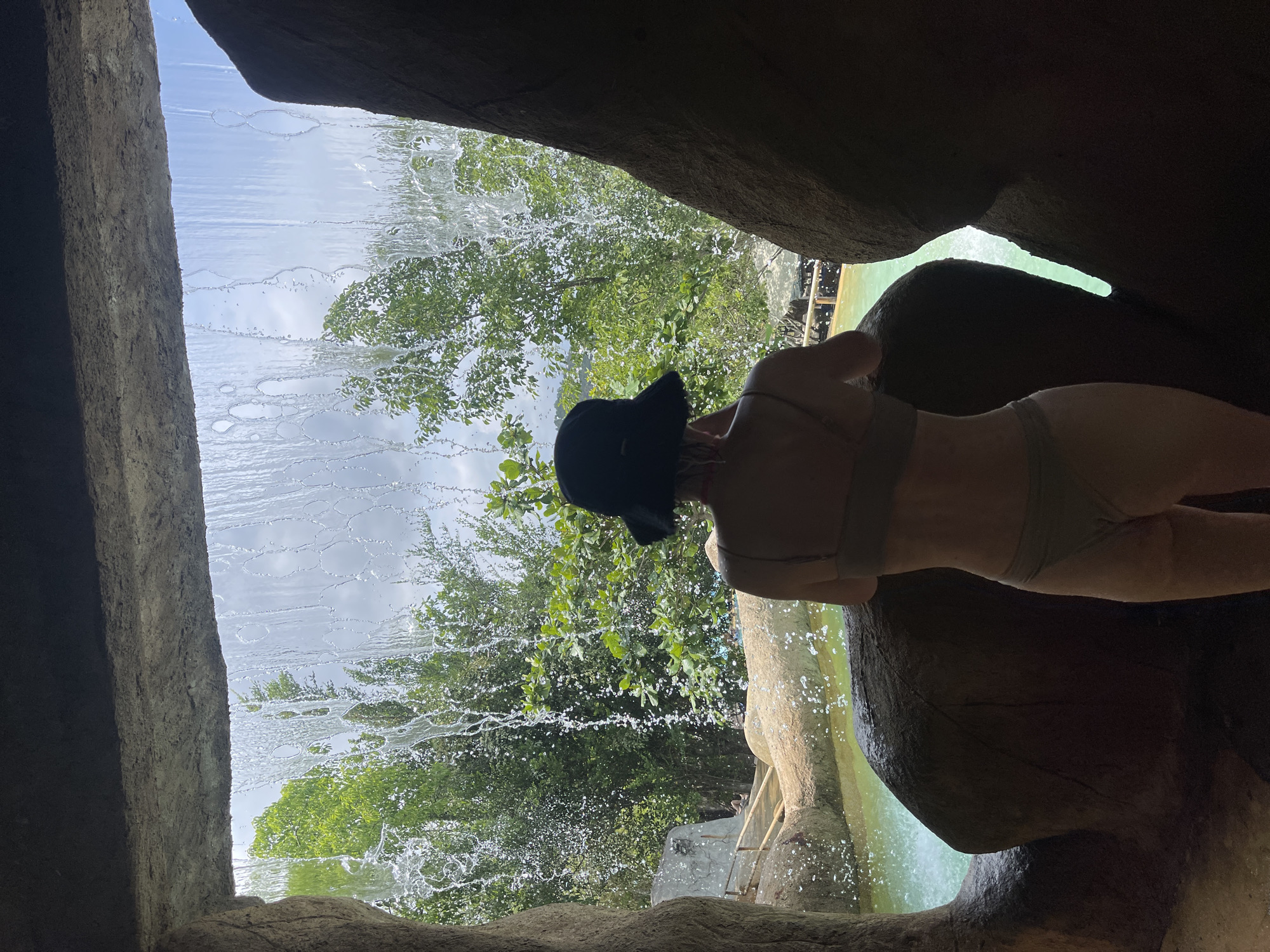 From within a rocky cave structure, a person is viewed from behind, wearing a bikini and a dark hat. The person stands near a waterfall, which is a prominent feature of the location. Outside the cave, lush green trees and foliage are visible. The scene is bathed in natural light. The location is in Tây Nha Trang, Vietnam.