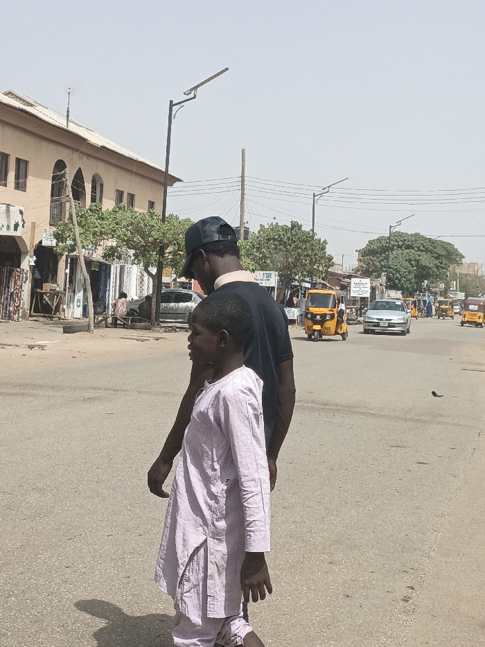 The image depicts an urban street scene in Hotoro South, Nigeria, under clear, bright daylight. In the immediate foreground, two individuals, both appearing to be male, walk away from the viewer on a gray asphalt road. The taller individual wears a dark t-shirt and a black baseball cap. The shorter individual, a child, walks slightly ahead and to the left, dressed in a light-colored, possibly pale lavender, long-sleeved tunic and trousers. Distinct shadows are cast on the sunlit pavement.
To the left of the road, a multi-story building with light-brown facades and arched windows stands, alongside street-level stalls displaying textiles and other goods. A discarded black tire rests near the curb. The street is lined with green, leafy trees and equipped with black streetlights featuring solar panels. Overhead utility wires are visible.
The mid-ground and background reveal an active urban thoroughfare. Multiple yellow motorized tricycles (keke napep) are present, some moving, some potentially parked. A silver sedan drives towards the viewer in the middle distance. Other vehicles and indistinct pedestrians are visible further down the road, contributing to the busy street environment.
