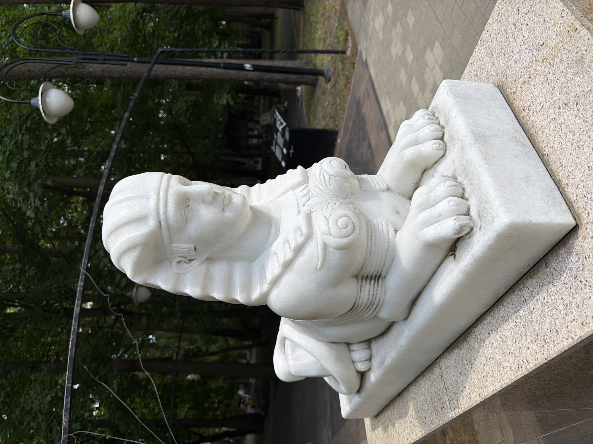 A white marble statue of a sphinx is centrally positioned outdoors in Svetlogorsk, Russia. The sphinx is depicted in a recumbent posture with its front paws extended forward. It features the head of a woman wearing a nemes-style headdress, with discernible facial features including eyes, nose, and closed lips. The female torso exhibits stylized breast detailing and a decorative, scroll-like pattern across the chest. The statue rests on a rectangular white marble base, which is set upon a larger, textured plinth composed of light brown and beige speckled granite. The background consists of a dense canopy of dark green deciduous trees. Multiple black metal lampposts with white, tear-drop shaped luminaires are visible, illuminating a paved pathway situated to the right and rear of the statue. In the distant background, blurred human figures and dark structures, including a prominent black information board with white text, are discernible. The scene suggests a public park or walkway environment under natural daylight.