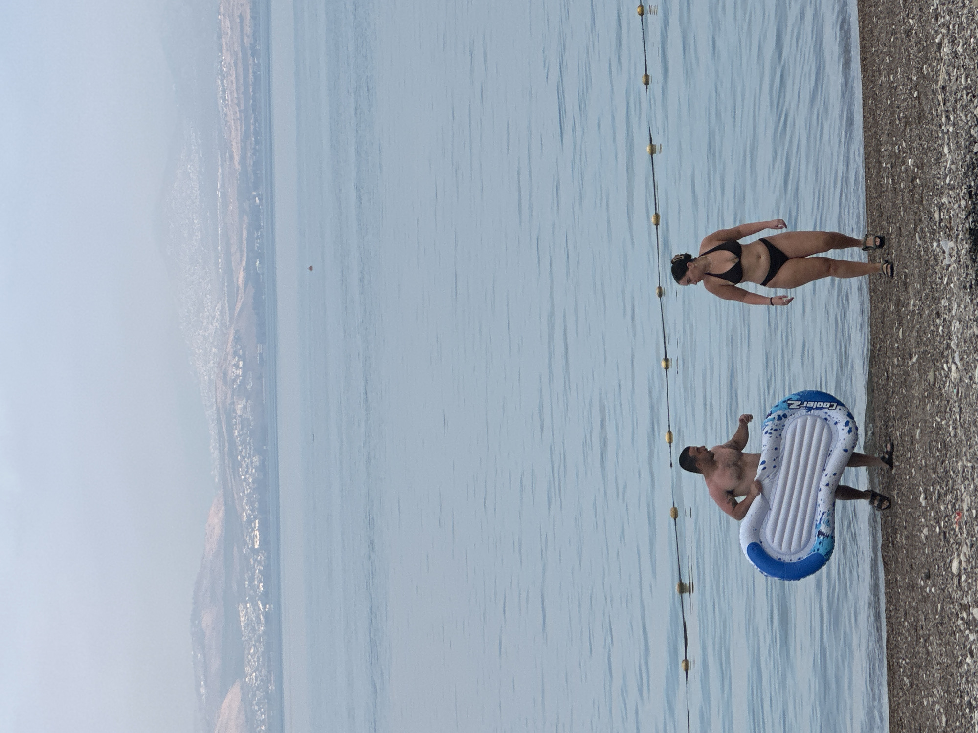 The image captures a serene beach scene in Emek HaYarden Regional Council, Israel, on a hazy day. A man, shirtless and wearing sandals, carries a blue and white inflatable raft along the pebbled shore. Facing him is a woman in a black bikini and sandals, walking towards the water. In the background, the calm, blue expanse of the Sea of Galilee stretches towards distant, hazy hills dotted with a town. A line of yellow buoys marks a boundary in the water. The overall atmosphere is one of leisure and relaxation by the water.