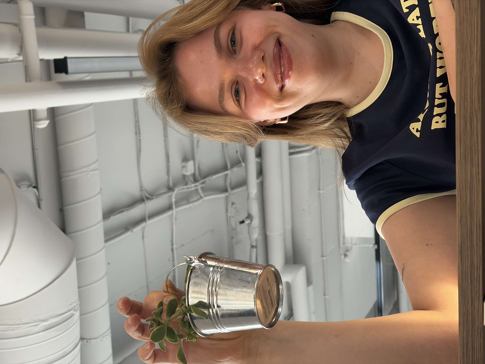 A fair-skinned woman with blonde hair, wearing a navy blue T-shirt with tan trim, is holding a small galvanized metal bucket with a plant in it. She is smiling at the camera. The background appears to be a white ceiling with exposed pipes and ducts. It is impossible to confirm that the image was taken in Бараўлянскі сельскі Савет, Belarus, from the given information.