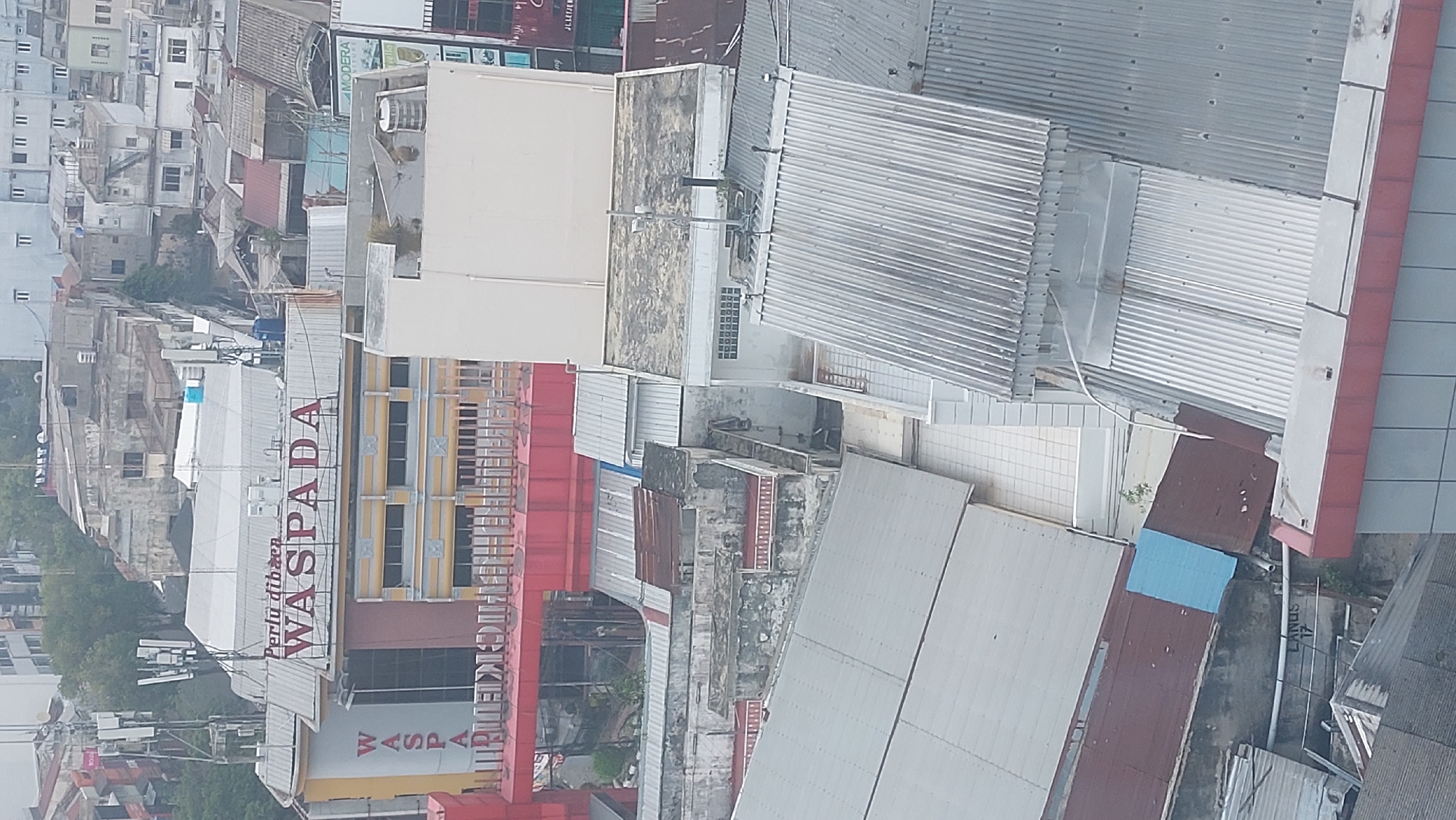The image shows an aerial view of a densely built urban area in the city of Medan, Indonesia. The foreground features several buildings with various roof types, including corrugated metal and flat roofs, some showing signs of wear and tear. Dominating the mid-ground is a building with a prominent red sign displaying "WASPADA" in large white letters. Below this sign, the building has a yellow and grey facade with multiple windows. A red architectural element, possibly an awning or a bridge, extends across the front of this building. In the background, a cluster of older, dilapidated buildings with weathered facades and a mix of roof materials can be seen, interspersed with trees and antennas. The overall impression is of a bustling city with a blend of older and newer structures.
