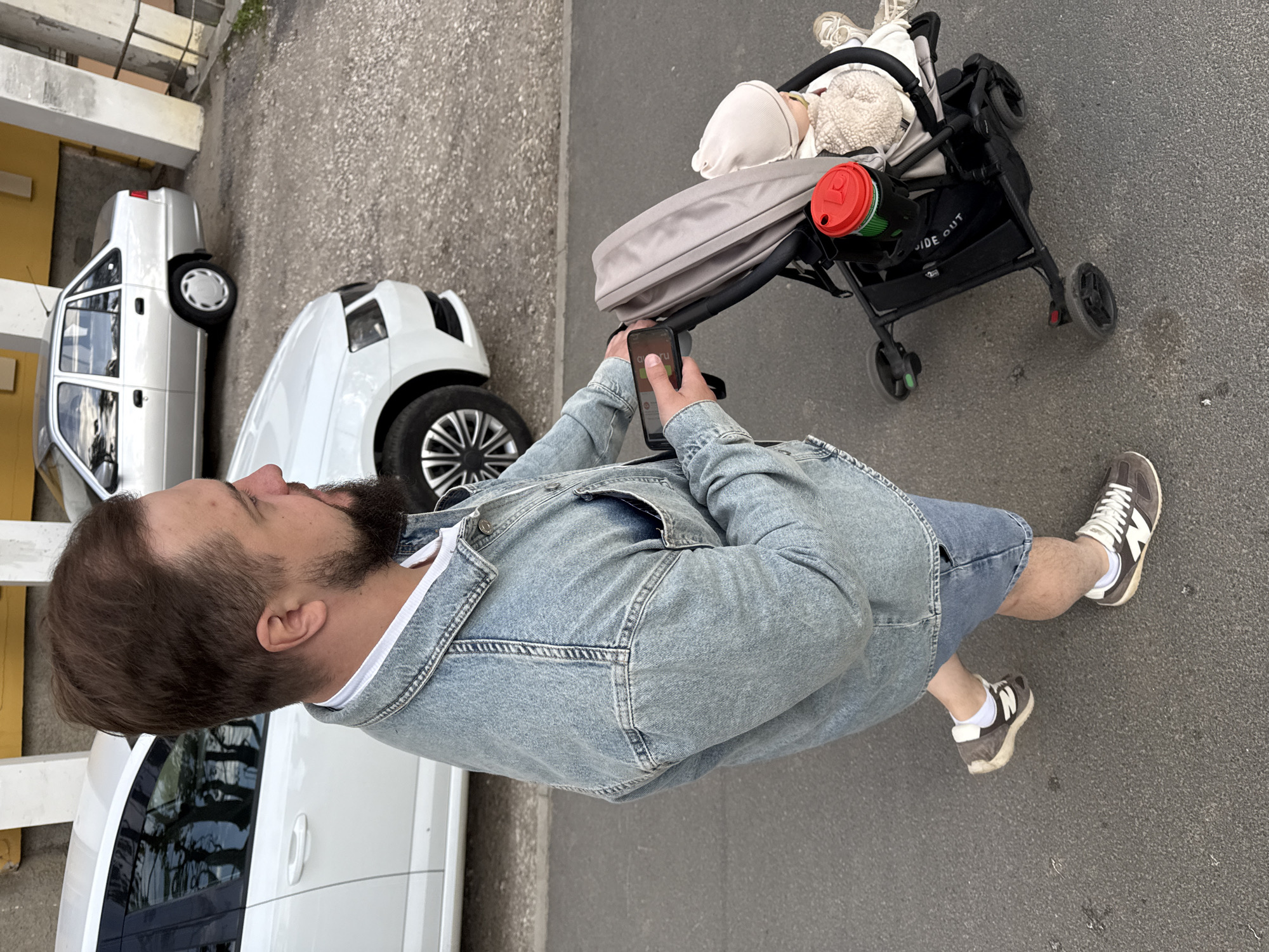 A man stands on asphalt next to a baby stroller and a row of parked cars. He is fair-skinned with a dark beard and hair, and he is looking at a phone in his hands. He wears a denim jacket, a white t-shirt, and blue shorts. On his feet are grey sneakers with white socks.
The stroller is black with a grey hood. Inside, a baby is wearing light-colored clothes. A red and green thermos is in the stroller's side pocket. The stroller is on the right side of the frame.
To the left are two white cars. A third car partially visible in the lower-left corner. The location has a yellow building in the background.
The image was taken outside in an urban area, likely in Vladimir, Russia.