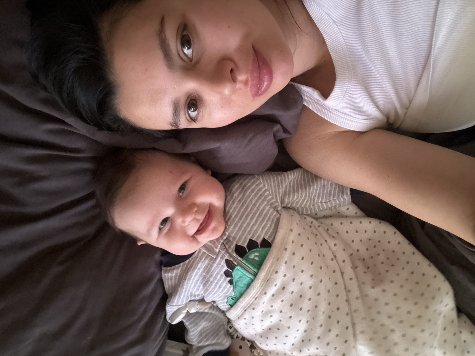 A woman and a baby are lying on a bed. The woman has dark hair and is wearing a white shirt. She is looking at the camera. The baby is wearing a striped onesie and a polka-dot blanket. The baby is smiling. The bed has brown sheets. The location is unknown.
