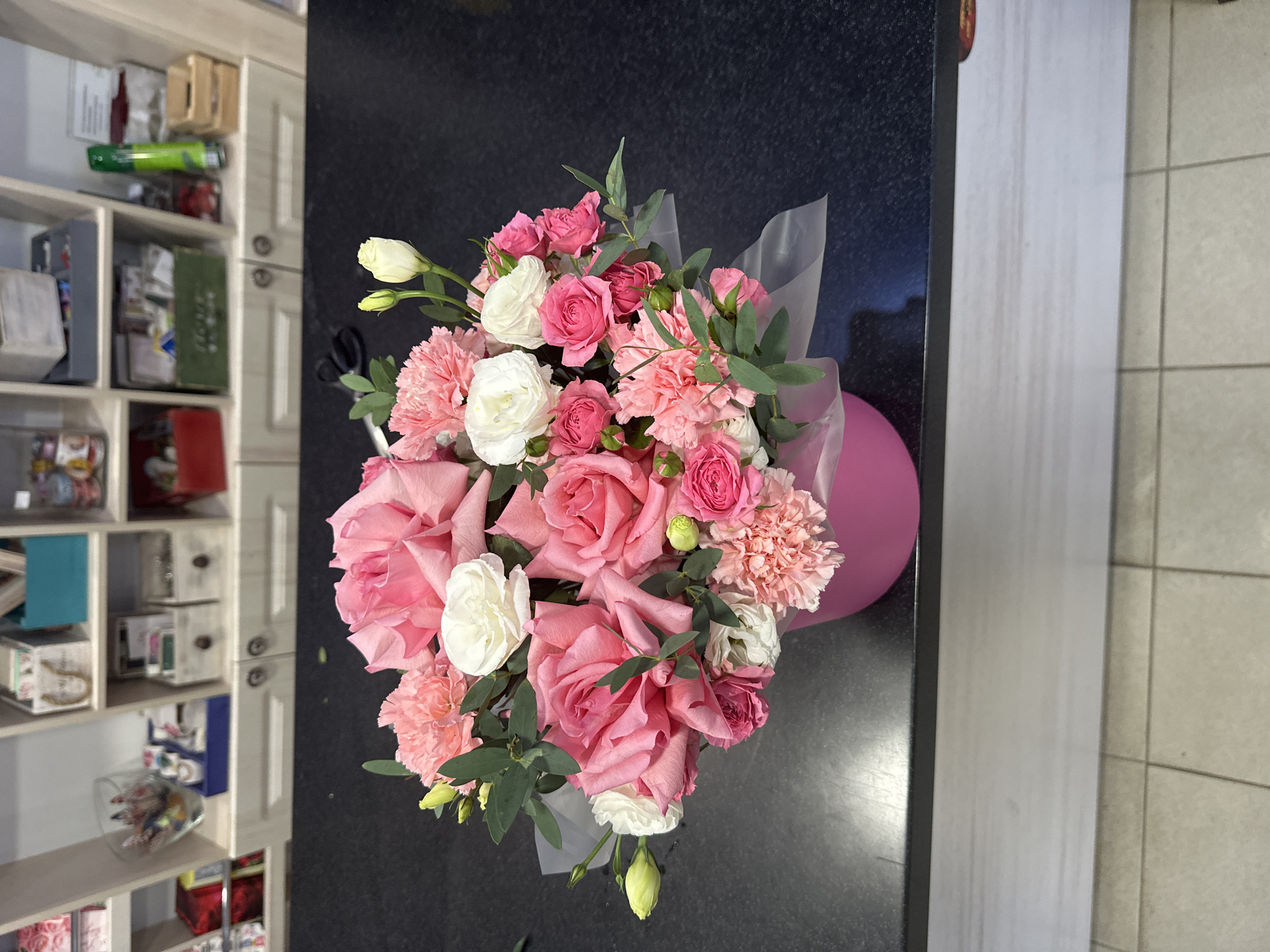 A pink cylindrical flower box containing a bouquet of pink roses, white ranunculus, and pink carnations sits on a dark, reflective surface, likely a countertop or table. The background reveals shelves filled with various items, including decorative boxes, jars, and what appear to be craft or stationery supplies. A pair of scissors rests on the dark surface to the left of the flower box. The overall scene suggests a retail or workshop environment, possibly a flower shop or a craft store, in Khanty-Mansiysk, Russia. The lighting is bright and appears to be artificial, casting subtle shadows.