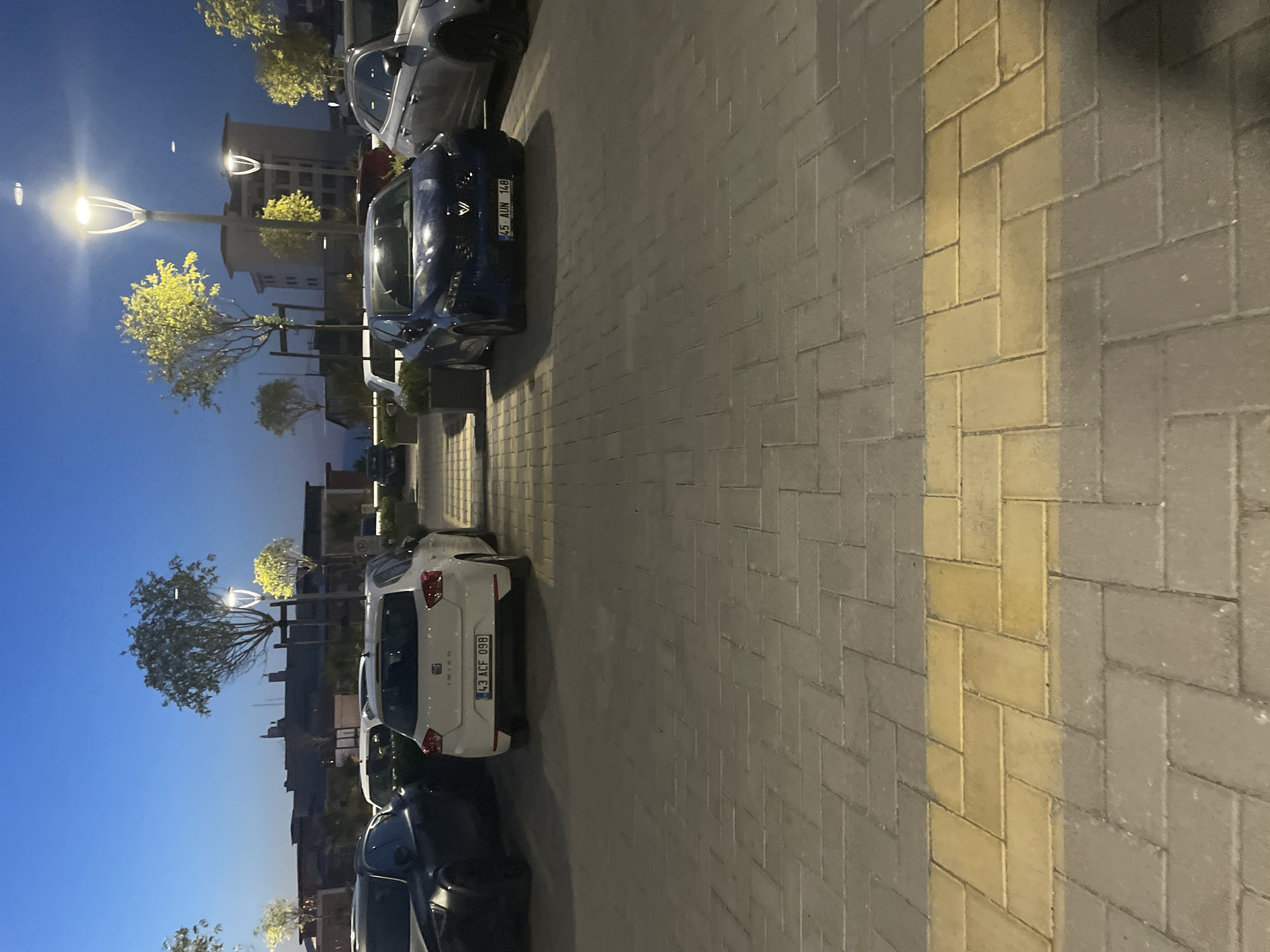 The image depicts a parking lot at dusk in Yunus Emre, Turkey, likely near a commercial area given the buildings in the background and the presence of streetlights. Several cars are parked, including a white SEAT Ibiza with license plate "43 ACF 09B" in the foreground, a dark blue car with license plate "45 AUN 148", and a silver car to its right. A dark-colored car is partially visible on the left. The parking lot is paved with interlocking bricks, with a distinct yellow brick pattern running horizontally in the foreground. Modern streetlights with illuminated, curved fixtures are spaced throughout the area, casting a warm glow on the scene. Trees with illuminated yellow foliage are planted near the streetlights. The sky is a deep twilight blue, indicating the transition from day to night. The overall environment suggests an urban or suburban setting during evening hours.