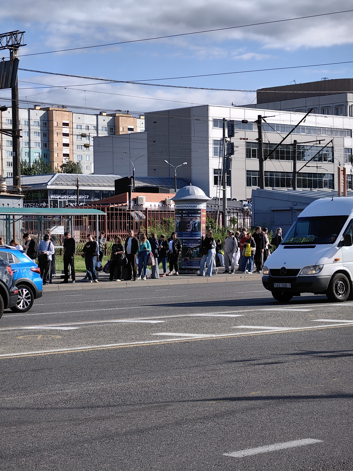 This media file captures a street scene in Minsk, Belarus, likely during daytime with a clear sky and scattered clouds. The foreground features a busy asphalt road with white lane markings, and a white van is driving on the right side of the road. To the left, a blue car is partially visible.
The main focus is a group of people waiting at a bus stop. They are standing on the sidewalk behind a metal fence, and many are looking at their phones. The bus stop itself has a clear roof and side panels. Behind the bus stop, there are several buildings. One building has "АПТЕКА" (Pharmacy) and "МЕДИЦИНСКИЙ ЦЕНТР" (Medical Center) written on it in large letters, suggesting this is a commercial area. Further in the background, there are residential buildings with visible windows. Overhead, power lines crisscross the sky.
The atmosphere appears to be one of everyday life, with people waiting for public transportation. There are no strong indicators of specific emotions, but the presence of a crowd suggests a common waiting period. The visible text provides specific location context within Minsk, related to businesses and advertising. The overall impression is a typical urban bus stop scene.