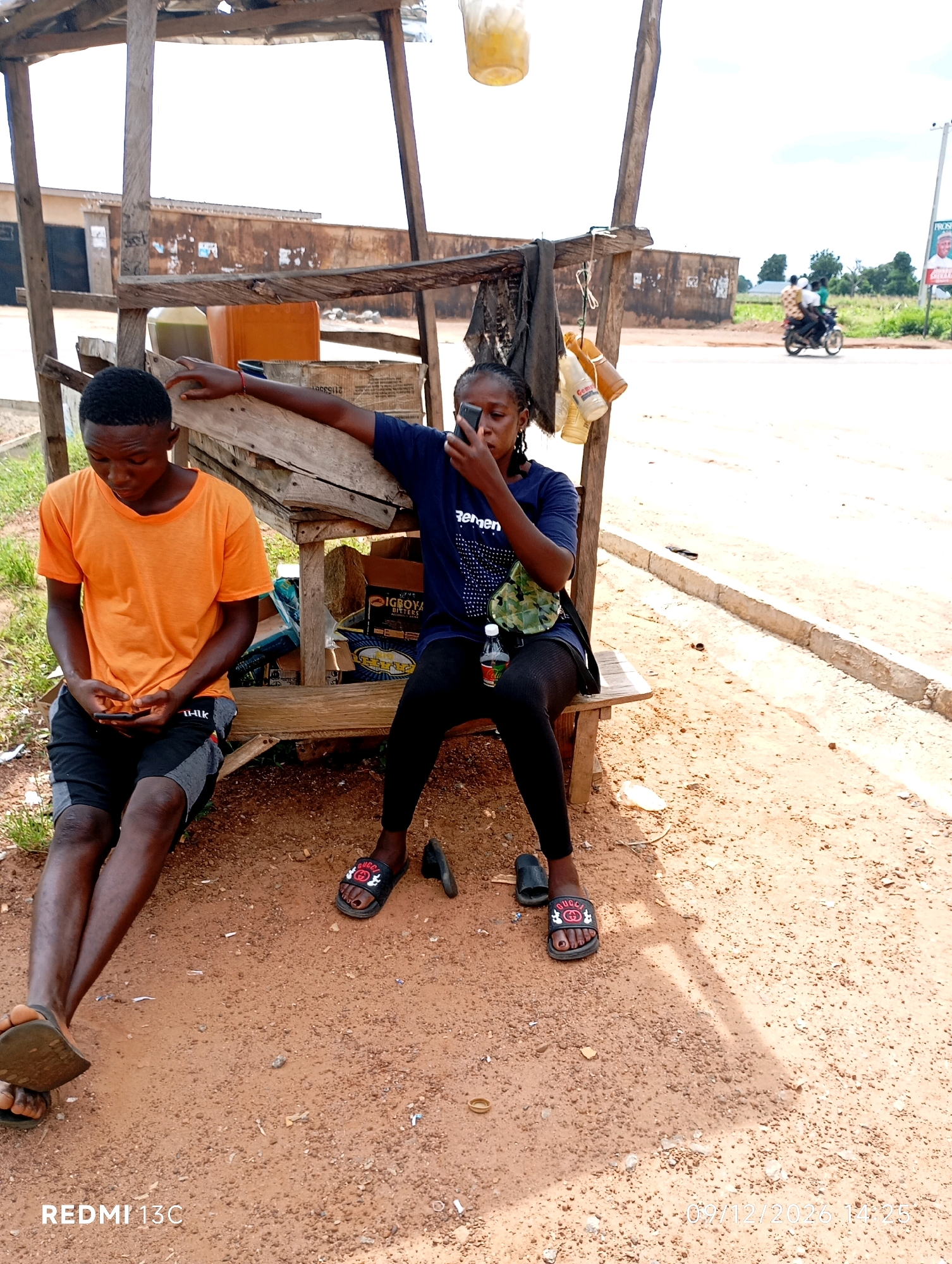 In Shendam, Nigeria, two young individuals are seated at an outdoor wooden stall. The stall is constructed from rough-hewn wood and appears weathered. The ground beneath them is reddish-brown soil, scattered with small stones and debris.
On the left, a young man wearing an orange t-shirt and black and gray shorts sits with his legs crossed. He holds a mobile phone in his hands and is looking down at it. He is wearing black sandals.
On the right, a young woman, also seated, wears a dark blue t-shirt with white lettering that appears to say "Zengey" and black leggings. She is holding a mobile phone up to her face, actively engaged with it. She has a camouflage-patterned bag slung across her shoulder and is wearing black slides with a red Gucci logo. Beside her, on the bench, is a bottle of water.
Behind the individuals, various items are visible within the stall. These include several bottles, some appearing to contain amber-colored liquid, possibly beverages or fuel. There are also cardboard boxes, one with visible text that might indicate a product name. A dark piece of fabric hangs from the stall's structure.
In the background, a road is visible with a concrete curb. Further in the distance, a billboard displays a portrait of a man with text in English. Several buildings with a rustic appearance line the road. A motorbike carrying three passengers is in motion on the road, heading away from the stall. The sky is bright and appears overcast or hazy. The overall scene depicts a moment of rest or waiting at a roadside stall.