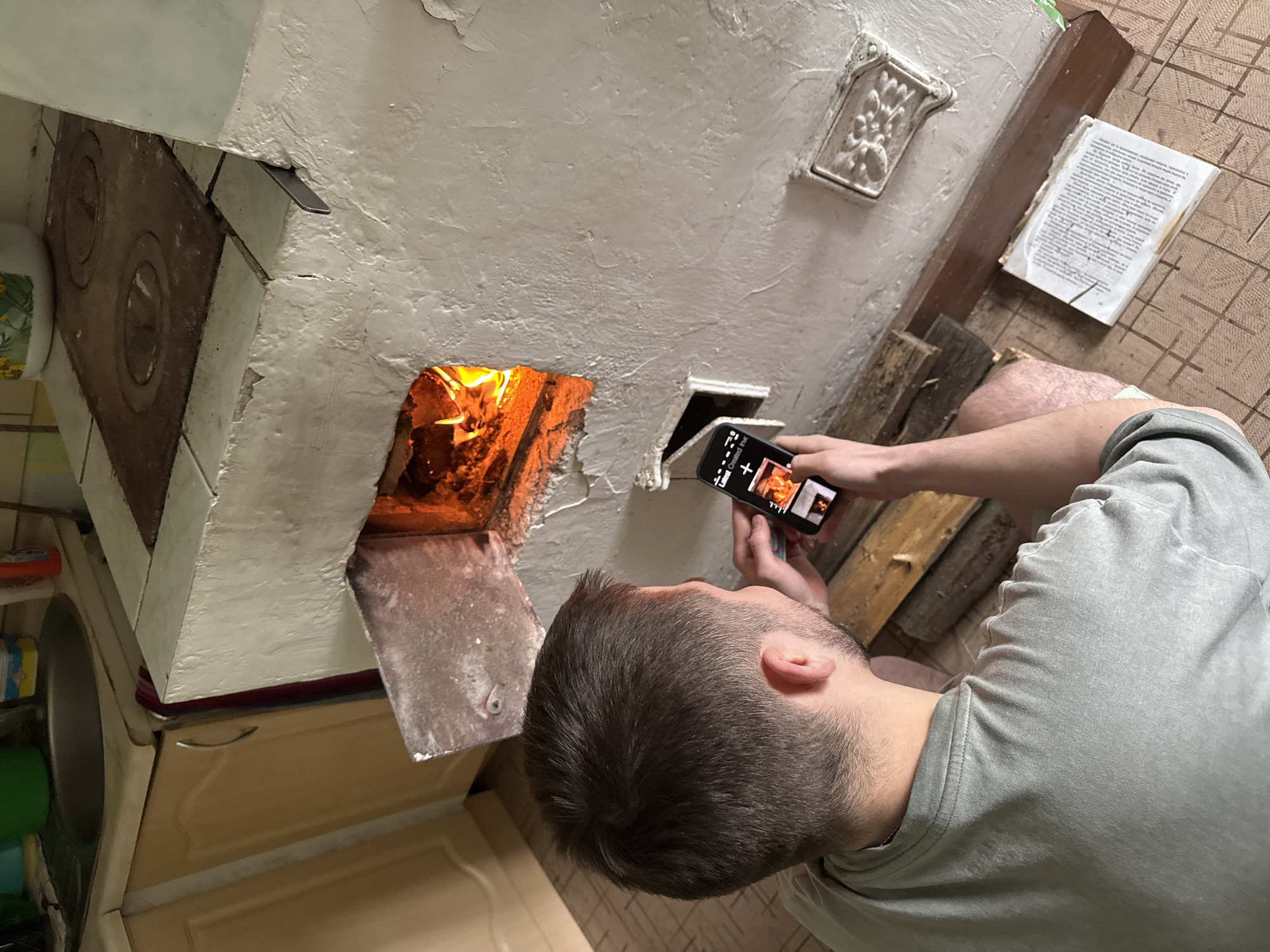 A person is photographing a fire in a white stove. The stove has a metal door, and is located in a room with a tiled floor and kitchen cabinets. There is wood stacked next to the stove and a piece of paper is on the floor. The person is wearing a gray shirt. The location might be Бородулино, Russia.