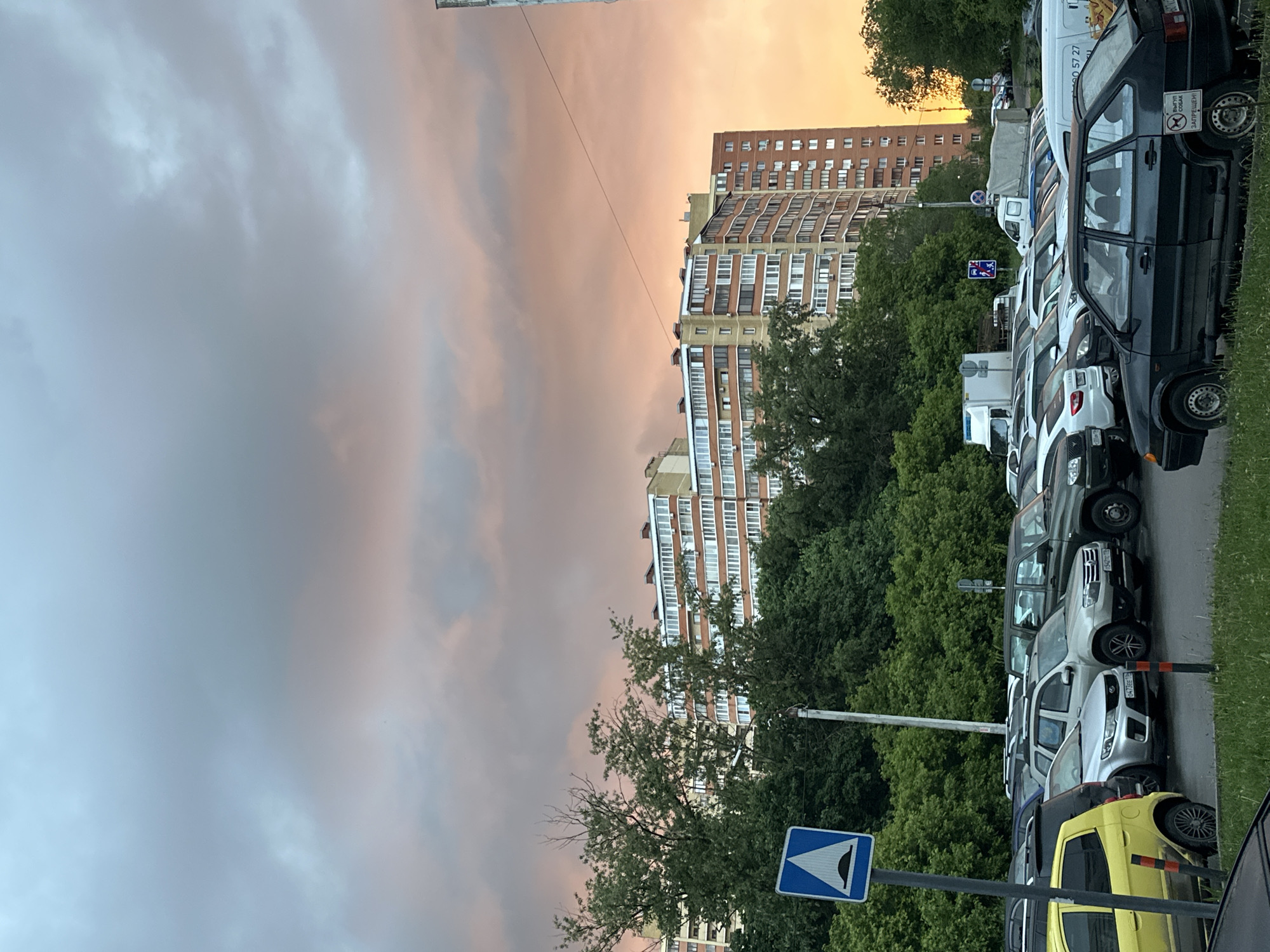 This media file captures an urban street scene, likely at either sunrise or sunset, given the dramatic coloration of the sky. The setting appears to be a residential area in Kazan, Russia, indicated by architectural styles and visible Cyrillic text.
The upper portion of the image is dominated by the sky, which displays a beautiful gradient of colors. On the left, clouds are a soft grey-blue, transitioning to warmer pink, orange, and yellowish hues towards the right, reflecting the low sun.
Below the sky, several multi-story residential buildings are visible. These structures feature numerous windows and balconies, characteristic of urban apartment blocks. A dense line of green trees stands between the buildings and the street, adding lush foliage to the landscape and partially obscuring the lower parts of the buildings.
Along the right side of the frame, a long row of diverse vehicles is parked. This includes various cars, SUVs, and vans, suggesting a residential parking area. The asphalt road surface appears somewhat damp. Towards the bottom right, a bright yellow car stands out among the other vehicles.
Notable details include:
- A blue road sign with a white triangular symbol, likely indicating an uneven road or speed bump, is visible on the left side, near the bottom.
- On the rear window of a black car, a white sticker with Cyrillic text reads: "ВЫГУЛ СОБАК ЗАПРЕЩЕН!" (Dog walking prohibited!).
- License plates on several cars show the regional code "16" (e.g., "043 ЕЕ 16", "047 ВЕ 16"), which corresponds to the Republic of Tatarstan, where Kazan is located.
- Part of a number, "D 57 27", is visible on the side of a white van.
The scene depicts a tranquil, everyday urban moment, devoid of people, with the focus on the natural beauty of the sky contrasted with the man-made environment of buildings and parked cars.
