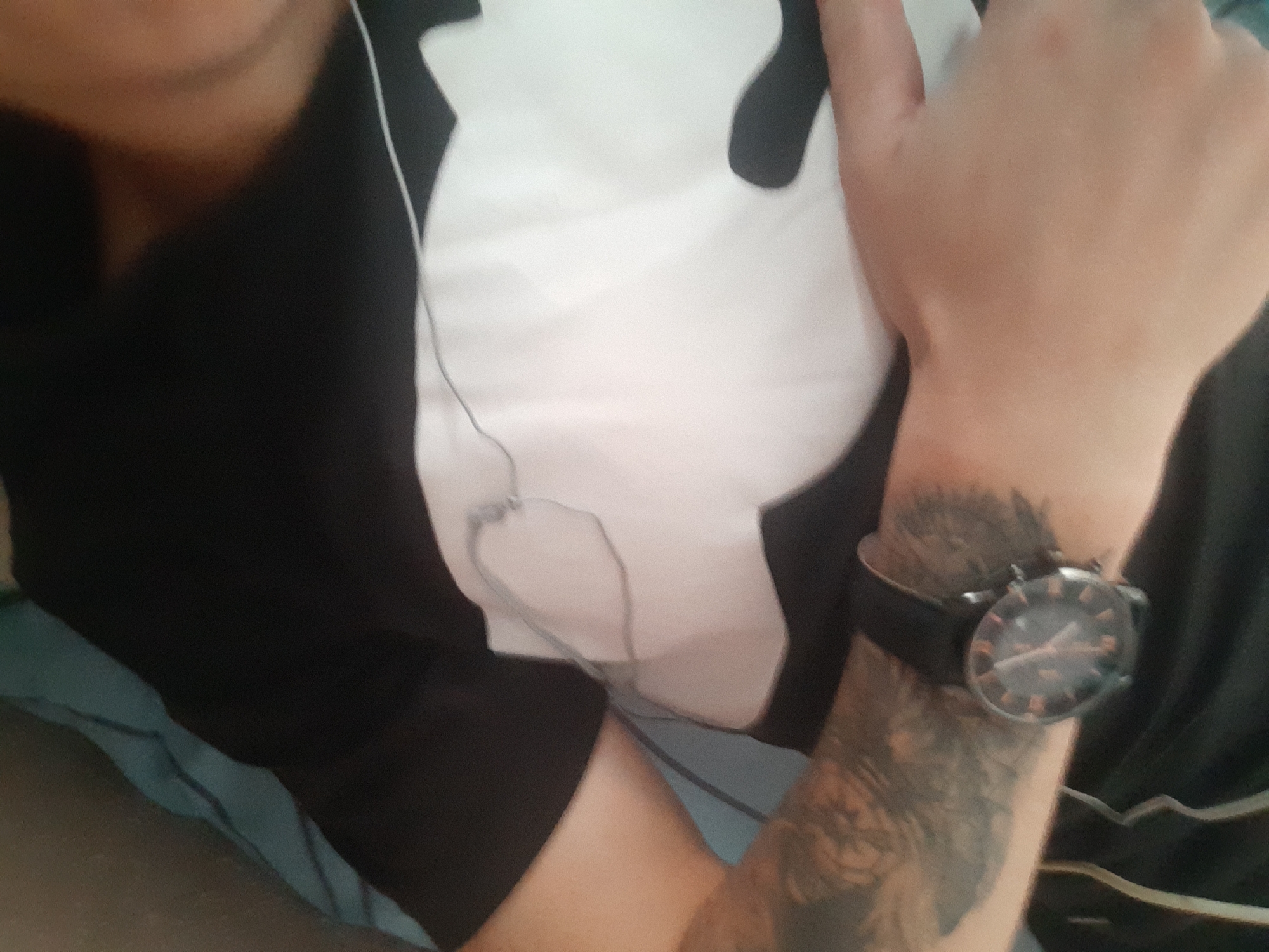 The image shows a close-up view of a person's arm and torso, wearing a black t-shirt with a large white graphic. A wristwatch with a dark band and an orange-accented face is visible on the left wrist. A tattoo covers a portion of the left arm. The person is lying on what appears to be a blue bed or sofa. The scene is indoors and there is no indication of the city of Cricova, Moldova.