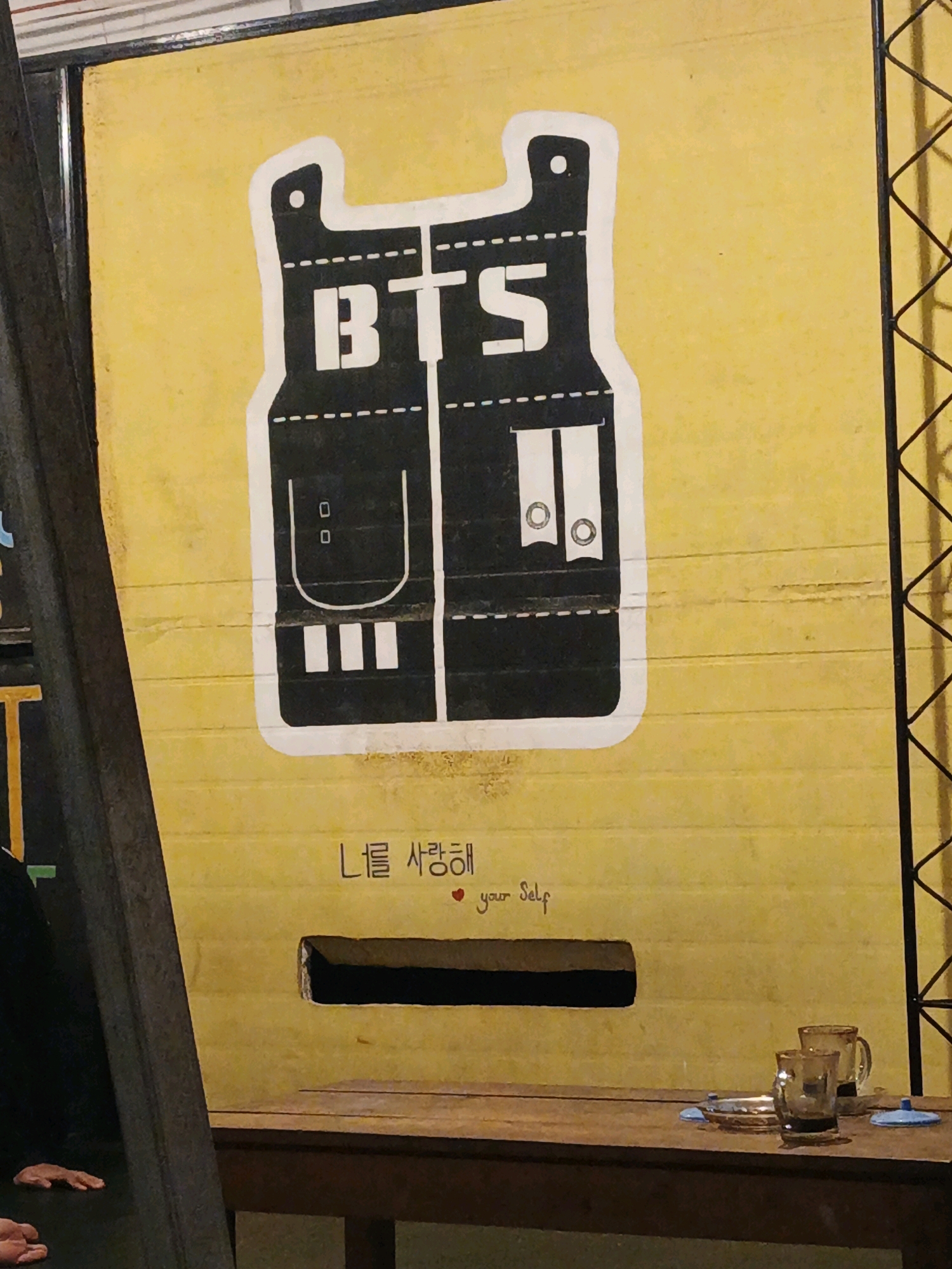 The image depicts a yellow wall with a large graphic of a black vest adorned with white lettering that spells out "BTS". Below the graphic, there is Korean text and the English phrase "love yourself" with a red heart. A rectangular opening is visible in the wall, through which a dark interior is seen. To the right of the opening, two glasses and a small dish are placed on a wooden surface. A black metal structure runs along the right edge of the image, and a dark vertical beam is present on the left. The overall scene suggests a creative installation, possibly in a public space like a cafe or art exhibit, within the city of Ketegan, Indonesia.
