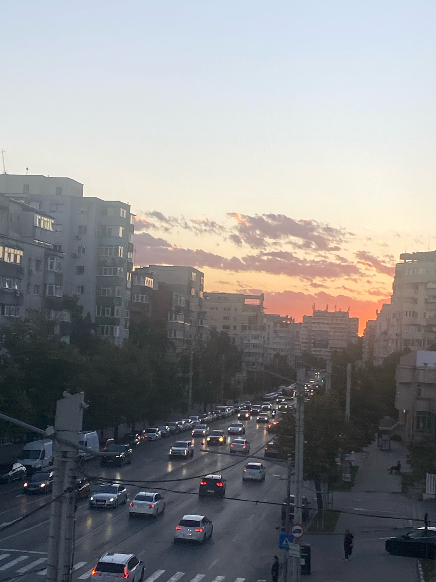 The image depicts a street scene in Iasi, Romania, during a sunset. The sky is filled with clouds tinged with orange and pink hues, indicating the sun is low on the horizon. Tall, apartment buildings line both sides of the street, their facades a mix of concrete and balconies. A multi-lane road runs through the center of the image, congested with traffic. Numerous cars, including sedans and SUVs, are visible on the road, some with their headlights on. A white van is parked on the left side of the street. Trees and other greenery are present along the roadside and between buildings. Pedestrians are sparsely visible on the sidewalks and near crosswalks. Electrical poles and wires are prominent in the foreground, adding to the urban environment. The overall impression is a busy city street at dusk with a vibrant sky.