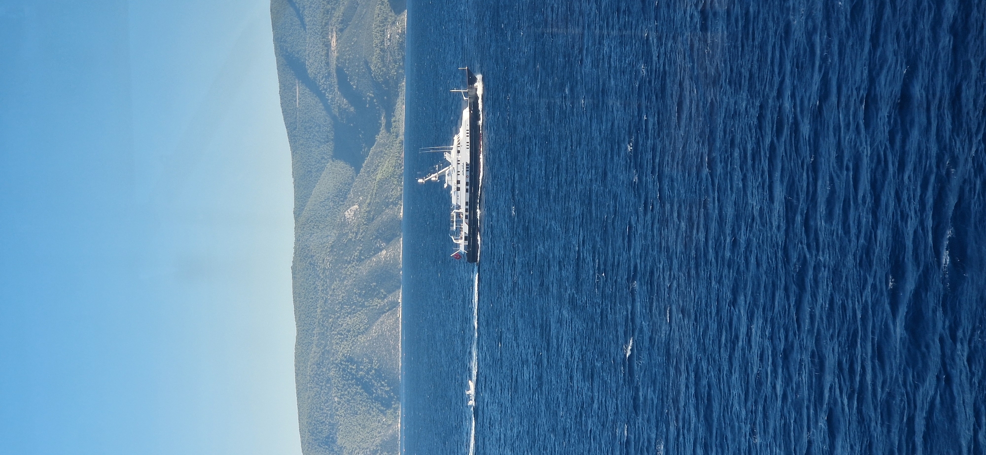 A large white yacht with a dark hull sails on a deep blue, choppy sea under a clear, pale blue sky. The yacht is moving from left to right. A smaller white boat is positioned to the left and slightly ahead of the larger yacht, also moving to the right. In the background, a densely vegetated, mountainous coastline stretches across the horizon. The overall scene depicts a sunny day at sea with vessels navigating near a natural landmass.