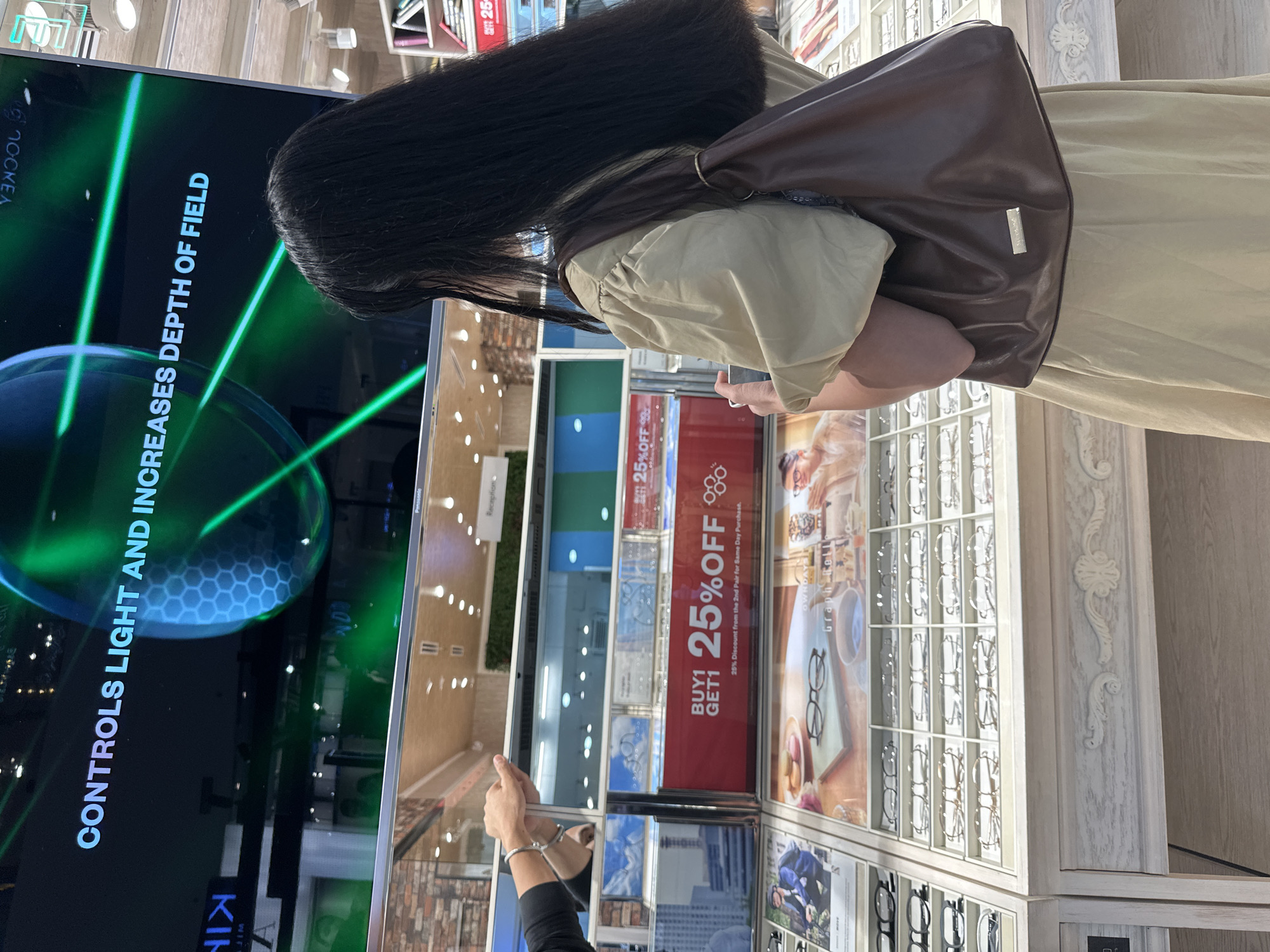 The image captures a young woman with long black hair, wearing a light-colored dress and a brown shoulder bag, standing in what appears to be an optical shop. She is looking at a large screen displaying text that reads "CONTROLS LIGHT AND INCREASES DEPTH OF FIELD" with green geometric graphics. In front of her, a display case exhibits various eyeglasses. Below the screen, a prominent red sign announces "BUY 1 GET 1 25% OFF". The environment suggests a retail setting, likely in Davao City, Philippines, given the context. The woman is holding her phone and seems to be contemplating a purchase. Other shoppers and store displays are visible in the background.