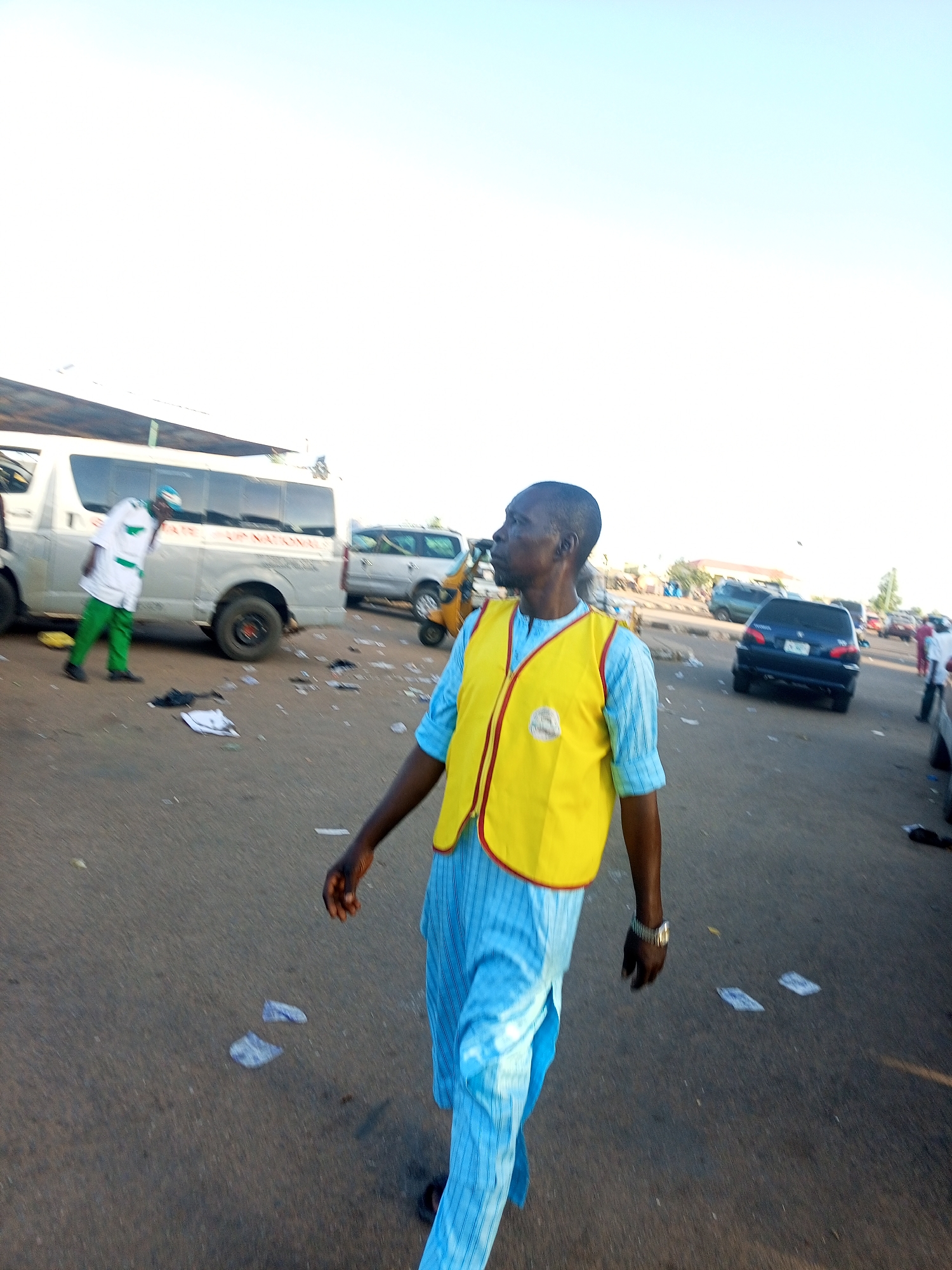 A man walks along a street littered with scraps of paper, wearing a light blue striped shirt and pants, and a bright yellow vest with red trim. He is glancing to his left. In the background, vehicles of various types are visible, including a van, a tricycle, and cars. Several people are standing around the vehicles. The sky is a pale blue. The location appears to be a busy street or public area in an urban environment, though the specific city and region are unclear.