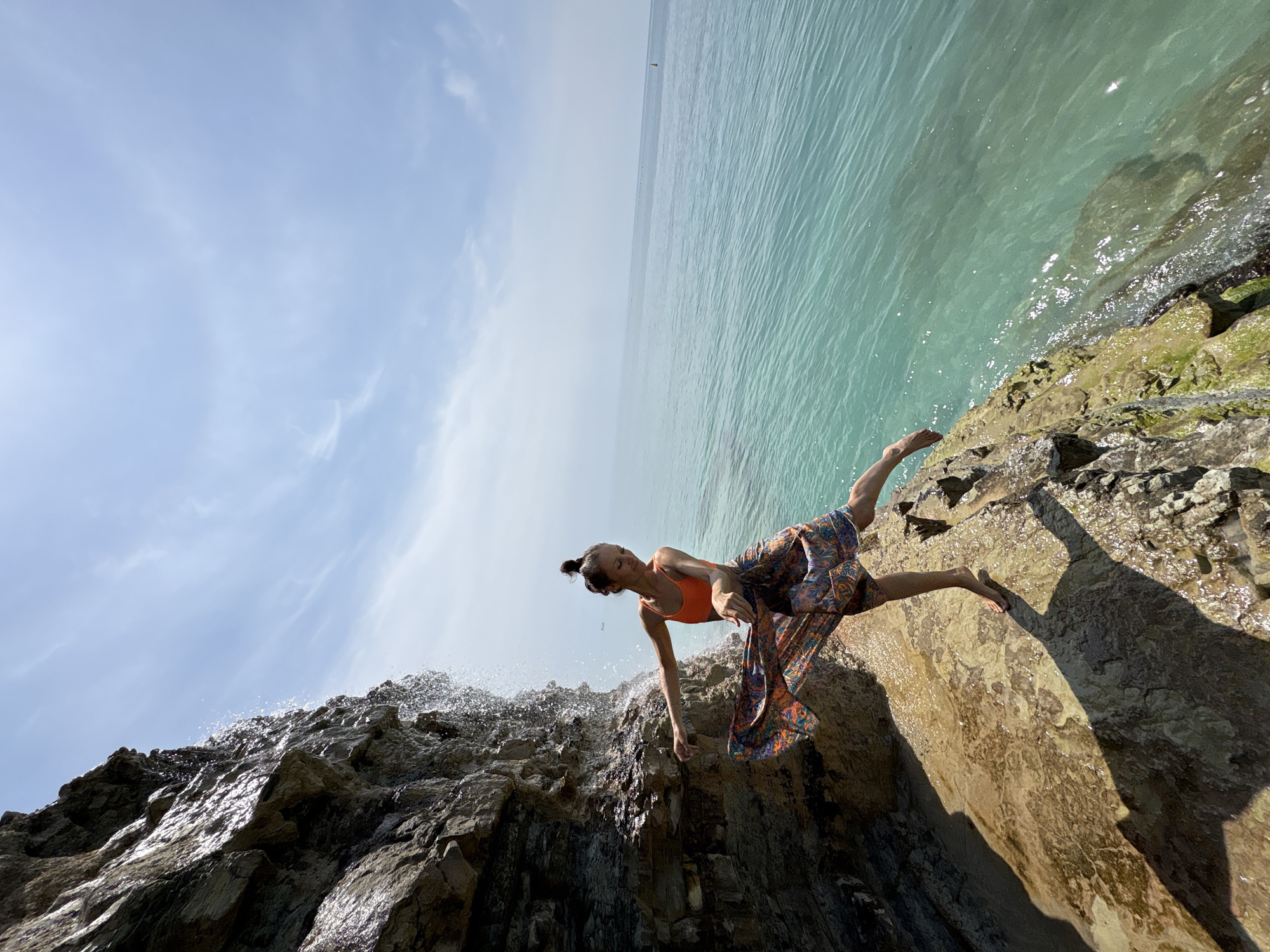 A woman is positioned at an extreme angle on a rocky cliffside. She wears an orange top and patterned skirt. The cliff descends into a body of water, likely the Black Sea. Water cascades over the rocks, indicating a waterfall or heavy splashing. The scene is oriented at a tilted perspective, showing the sky and sea horizon at an angle. The location is likely near Gelendzhik, Russia, a coastal area known for cliffs and beaches.