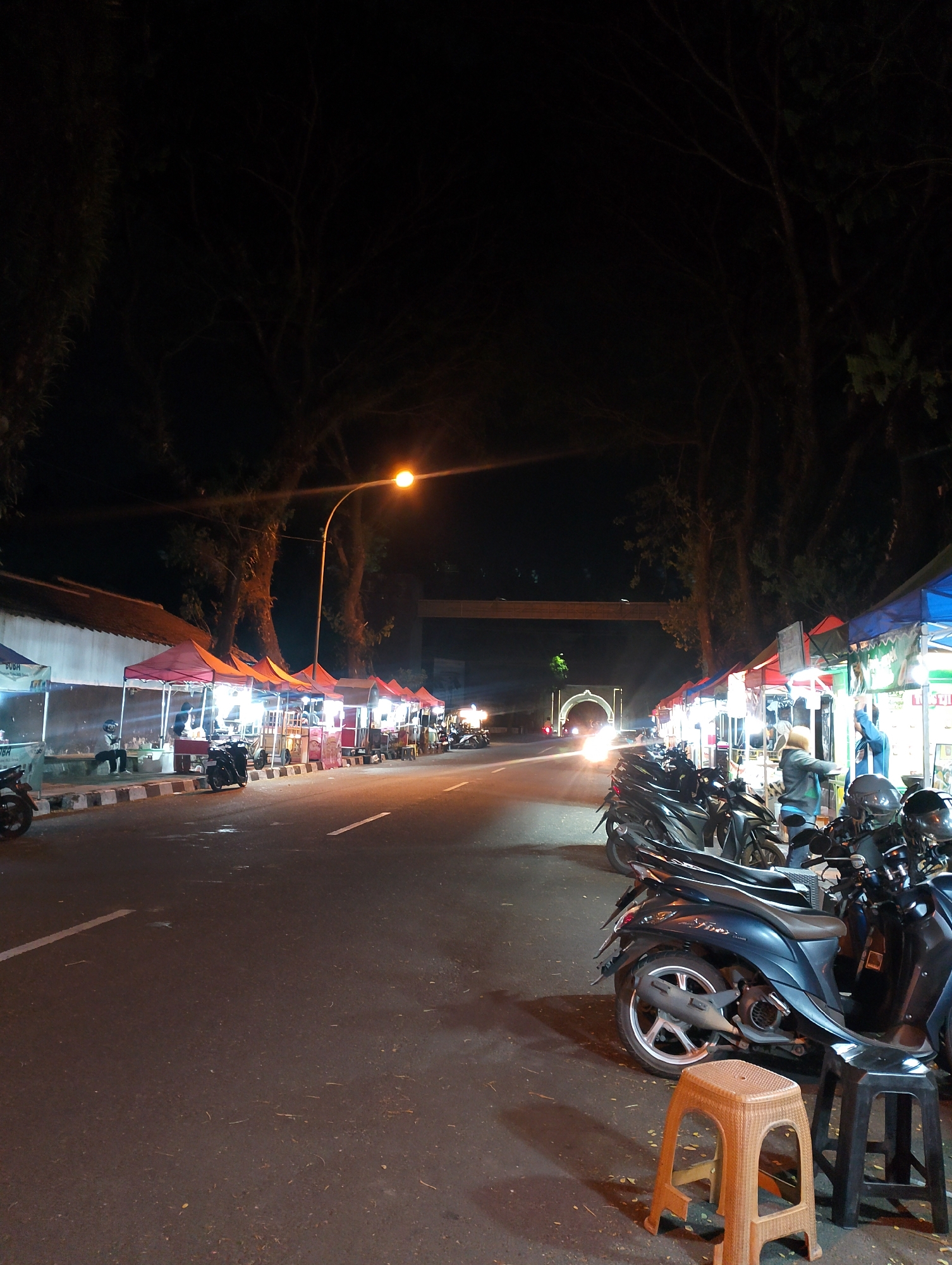 This is a nighttime, vertical, medium-shot photograph of a street scene in Magelang, Indonesia, likely featuring a night market. On the left side of the image, there is a row of market stalls with red and orange awnings, illuminated by artificial lights. Some of these stalls appear to be selling food. Several motorbikes are parked in front of the stalls. A person is visible sitting on a bench at one of the stalls.
The street itself is paved and appears wet in places, reflecting the light. On the right side of the street, there is another row of market stalls with blue and green awnings. Several motorbikes are parked in front of these stalls as well. Two people are visible standing at one of these stalls, one of whom is wearing a blue jacket and the other a grey hoodie. A few plastic stools are placed near the parked motorbikes.
In the background, large trees with bare branches line the street. Further in the distance, a brightly lit archway is visible, suggesting a landmark or entrance. The overall atmosphere is that of an active urban night market, with the glow of lights illuminating the food stalls and the surrounding street.