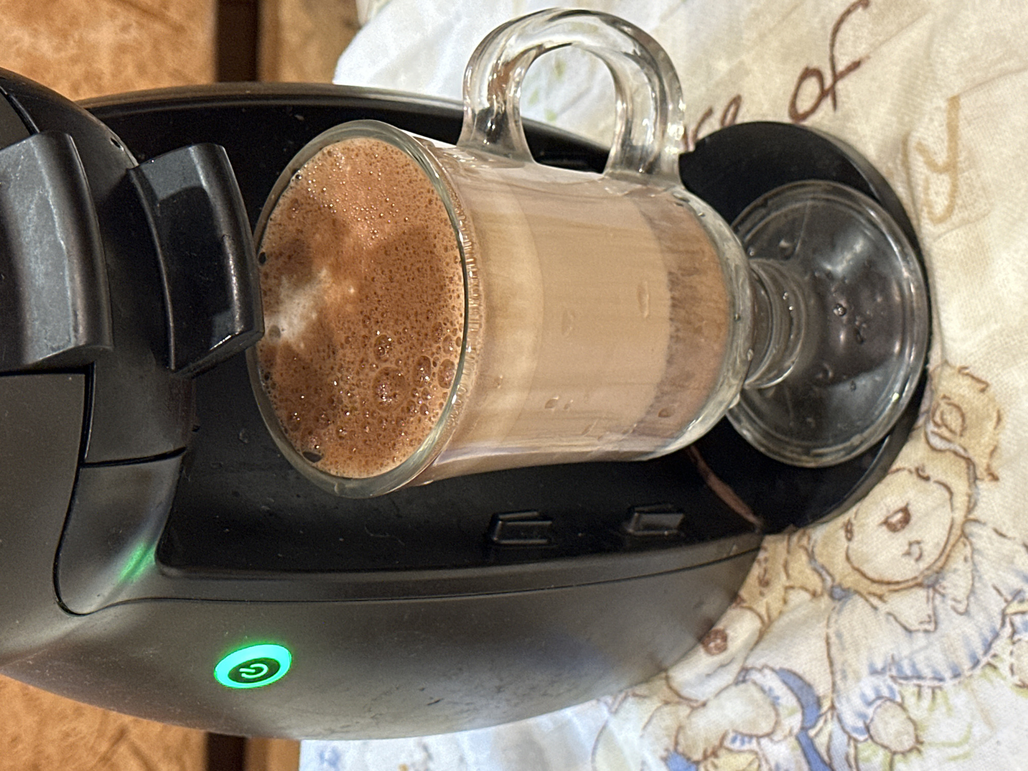 A close-up, high-angle shot focuses on a black coffee maker, its green power light illuminated. To the right, a clear glass mug filled with a frothy, chocolate-colored beverage sits on a dark base. The mug has a distinct handle and a stemmed foot. The coffee maker and mug are placed on a white fabric surface with a faded pattern of teddy bears and cursive writing. The background consists of a textured, wood-like surface. This image is not located in Nizhny Novgorod, Russia.