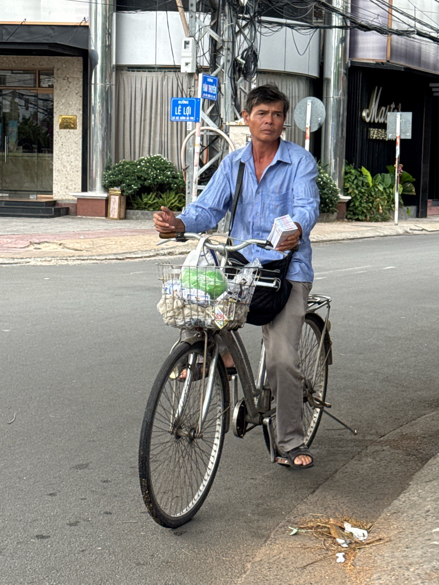 A middle-aged Vietnamese man is riding a bicycle on a paved city street. He is wearing a blue collared shirt and gray pants, with black sandals. His hands are on the handlebars, and he is holding a stack of what appears to be money in his right hand. The bicycle has a front basket containing some items, including a green plastic bag and a black bag. The street is lined with buildings, one of which has signage that reads "MASTER." There are also street signs visible, including one that reads "DUONG LE LOI," indicating the location is Le Loi Street in Nha Trang, Vietnam. The environment appears to be an urban commercial area with some greenery and overhead electrical wires. The man's expression is neutral.