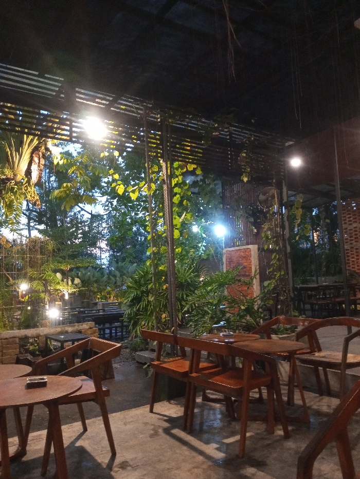 The image presents an unoccupied, semi-outdoor dining or cafe area at night, located in Pontianak, Indonesia. The scene features multiple wooden tables and chairs, illuminated by artificial overhead and ground-level lighting. In the foreground, a round wooden table holds a dark ashtray, surrounded by two wooden chairs with slatted backs. To the right, additional wooden tables and chairs are arranged. The environment is heavily integrated with lush tropical vegetation, including large-leafed plants, climbing vines, and a hanging fern on the left. A dark, slatted roof structure with metal supports covers the area. Paved ground, appearing dark and reflective, is visible throughout. A wall-mounted fan is situated on the right side of the frame, between structural elements and foliage. Further seating and a partial brick wall are visible in the background. No individuals are present in the image.