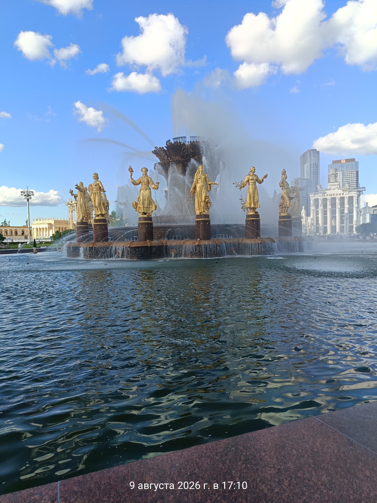 The image shows a large, ornate fountain with golden statues of women in classical attire. The fountain is in the foreground, with water spraying upwards and cascading down. In the background, there are modern buildings and a clear blue sky with white clouds. The overall scene depicts a public space, likely a park or square, in an urban environment. There are no people clearly visible in the image, but there is a suggestion of activity in the background. The date and time stamp "9 августа 2026 г. в 17:10" is visible in the bottom right corner of the image, suggesting it was taken on August 9, 2026, at 17:10.