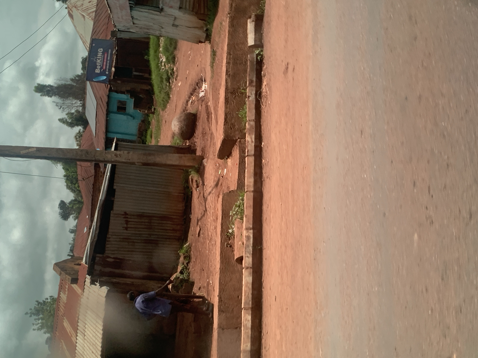 This image depicts a street scene in Hoss, Nigeria. In the foreground, a paved road with a reddish-brown dirt shoulder is visible. A concrete curb separates the road from the adjacent area. The middle ground reveals a dirt path with scattered debris and vegetation. A man, dressed in a blue shirt and dark pants, is sweeping the dirt path with a broom. Behind him, a corrugated metal structure with a red tin roof is partially visible, and further back, a light blue building with a small window stands. A large, dark sign with white lettering, reading "BetKING", is mounted on one of the structures. Several other buildings with tin roofs are present in the background, along with trees and a cloudy sky. The overall environment appears to be a mix of residential and commercial structures in a somewhat unkempt condition.