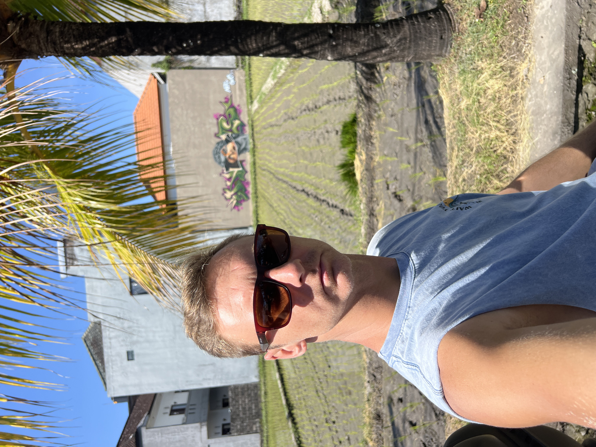 A medium shot selfie shows a Caucasian man with short blond hair and sunglasses in the foreground, wearing a light blue sleeveless shirt. He is positioned in front of a rice paddy field in Sukawati, Indonesia. Behind him, a wall with graffiti is visible, featuring abstract designs and a portrait. Further in the background, there are buildings and a palm tree. The sky is clear and blue.