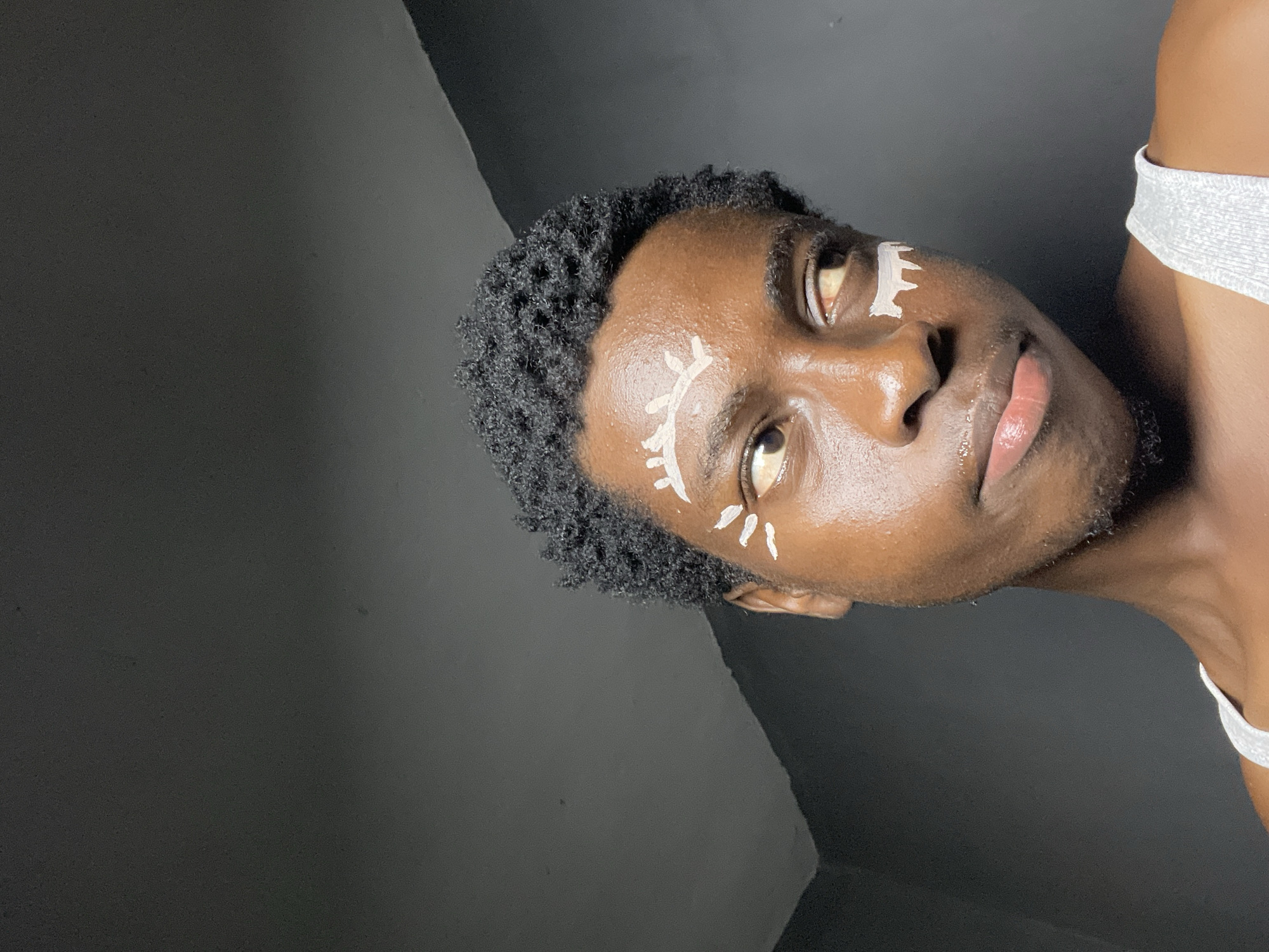 The image shows a close-up, head-and-shoulders portrait of a young Black person with dark, curly hair. They are looking off to the right side of the frame with their eyes slightly narrowed. White face paint is applied in decorative patterns around their eyes, resembling stylized eyelashes and a brow. They are wearing a white tank top, and their skin appears to have a dewy sheen. The background is dark and textured, suggesting an indoor setting, possibly a room with dark walls and a ceiling. The lighting is dim, creating shadows that highlight the contours of their face. The overall impression is one of artistic expression and self-portrayal. There is no visible indication of the location being Nkpor, Nigeria, or any urban environment.