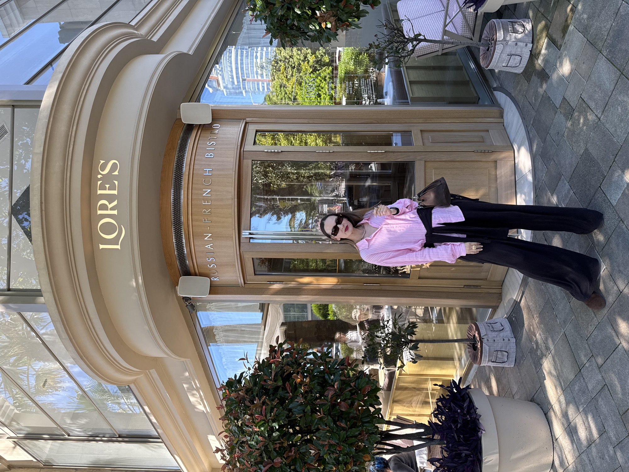 An outdoor, full-length shot captures a woman posing in front of "LORE'S RUSSIAN FRENCH BISTRO" in Sochi, Russia. The woman stands centrally, wearing a pink and white striped button-down shirt, black wide-leg pants, and dark sunglasses, with a dark brown handbag on her shoulder.
The restaurant features a grand, light-colored arched facade with ornate moldings, displaying "LORE'S" in prominent gold letters. To the right, "RUSSIAN FRENCH BISTRO" is written vertically on a wooden panel. The entrance consists of large glass doors and windows that reflect the bright, sunny sky, lush green trees, and modern buildings in the background. An outdoor speaker is mounted above the right side of the entrance.
The ground is paved with grey interlocking stones, indicating an outdoor pedestrian area. Several potted plants and decorative planters, one with a unique light-colored patterned fabric covering, are arranged near the entrance. The scene is illuminated by clear daylight, casting distinct shadows and suggesting good weather.