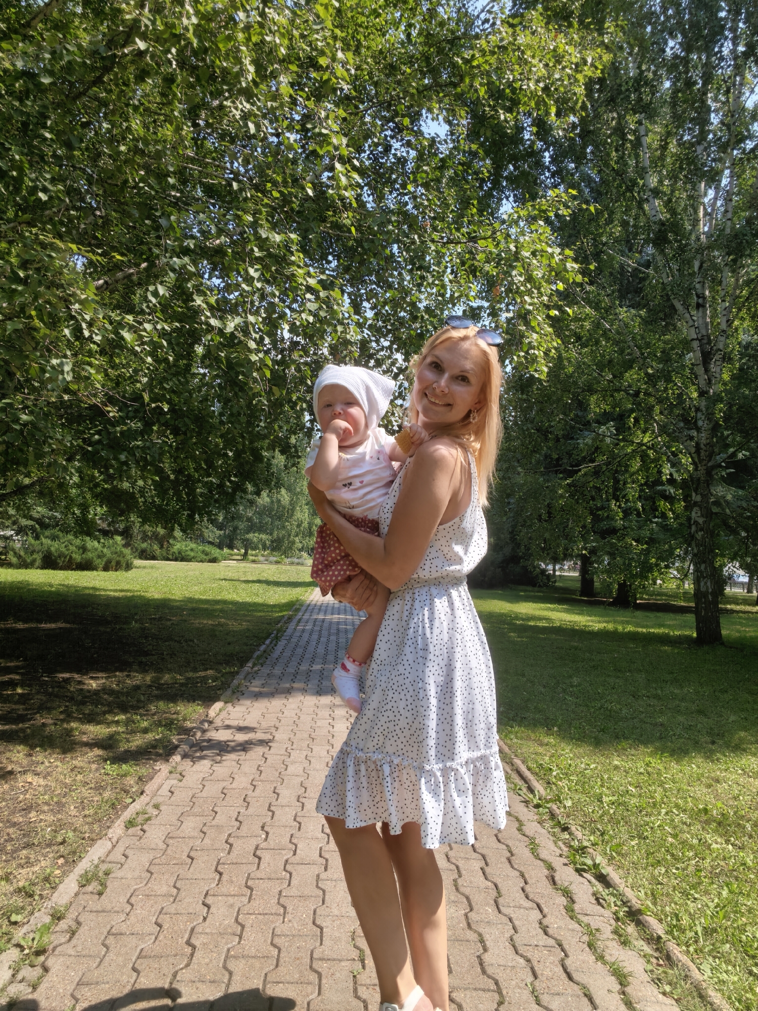 A woman with blonde hair, wearing sunglasses on her head and a white polka-dot dress, smiles at the camera while holding a baby. The baby, wearing a white head covering and a pink and white outfit, is looking away from the camera. They are standing on a paved walkway in a park with trees and green grass. The scene appears to be outdoors on a sunny day.