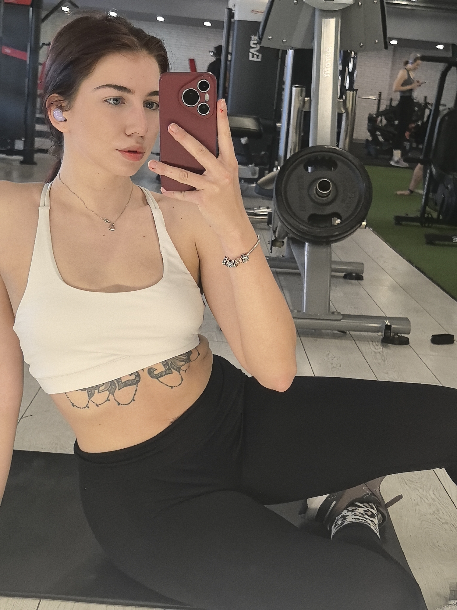 A fair-skinned young woman poses for a selfie indoors, likely in a gym. She wears a white sports bra, black leggings, and patterned socks with running shoes. A floral tattoo is visible on her lower abdomen. She holds a dark red smartphone in her right hand, obscuring part of her face. An earbud is visible in her left ear. She wears a necklace and a charm bracelet.
Behind her is gym equipment, including a weight bench with a barbell. Other gym-goers are faintly visible in the background.
The location is potentially a gym in Stara Zagora, Bulgaria.