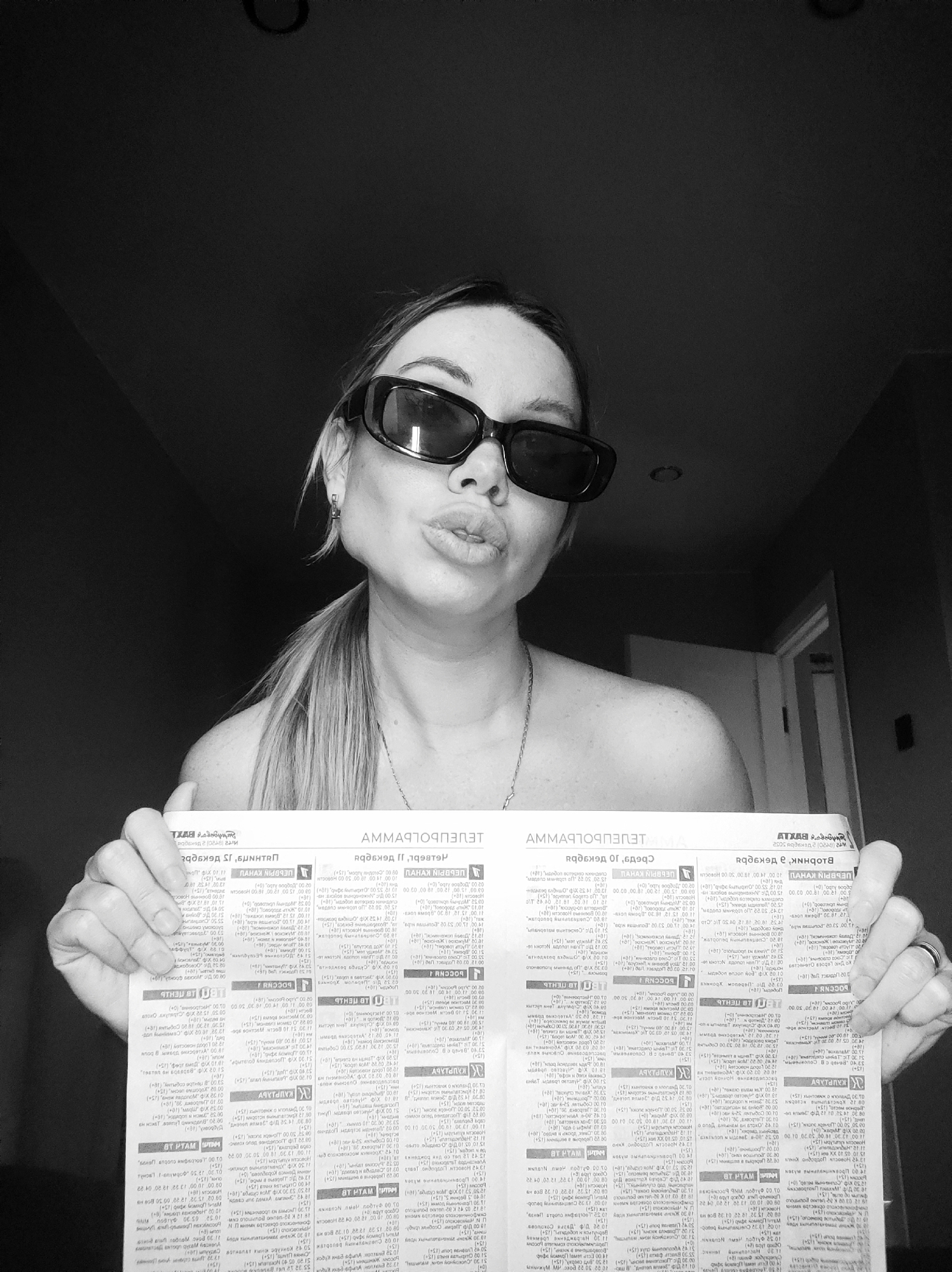 A black and white photograph captures a woman indoors, posing for the camera. She has light-colored hair styled in a ponytail and wears dark, rectangular sunglasses. Her lips are pursed in a playful "kissy face" expression, and she looks directly at the viewer. She appears to be bare-shouldered, possibly shirtless or wearing a strapless top, and a delicate chain necklace is visible around her neck.
With both hands, she holds up a folded piece of paper, likely a newspaper or a television program guide, in front of her chest. The text on the paper is printed in Cyrillic and is mirrored, indicating it was either held reversed or the image was flipped horizontally. Prominently visible, though mirrored, is the word "ТЕЛЕПРОГРАММА" (Teleprogramma), which translates to "TV program" in Russian. Other discernible, mirrored Cyrillic words and phrases consistent with a TV schedule include "КАНАЛ" (Channel), "РОССИЯ" (Russia), "ВТ МАТЧ" (likely referring to a sports channel), and "КУЛЬТУРА" (Culture).
The background is dark and dimly lit, suggesting an interior setting in Votkinsk, Russia. A partial view of a ceiling light fixture can be seen at the top, and a doorframe or opening is visible on the right side of the image. The overall mood is casual and confident.