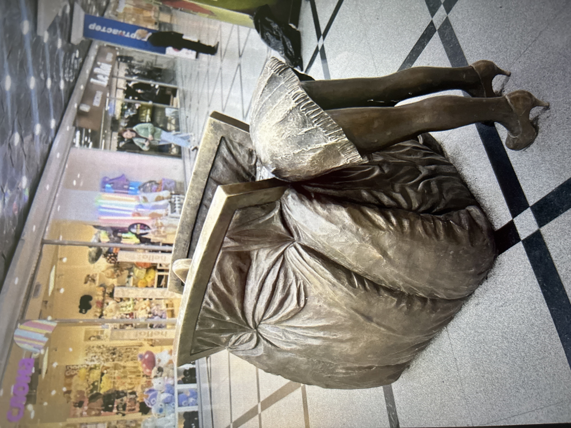 The image depicts a bronze sculpture of a woman's legs wearing high heels, emerging from a large, draped bag. The sculpture is situated on a checkered tiled floor within a shopping mall. In the background, storefronts are visible, displaying merchandise and signage, including what appears to be a store with the word "Le Roi" and another with "Картымастер" (Kartamaster). People are walking in the background, some appearing to be shoppers or employees. The overall impression is an art installation within a commercial public space. The presence of the Cyrillic signage suggests a location in Russia.