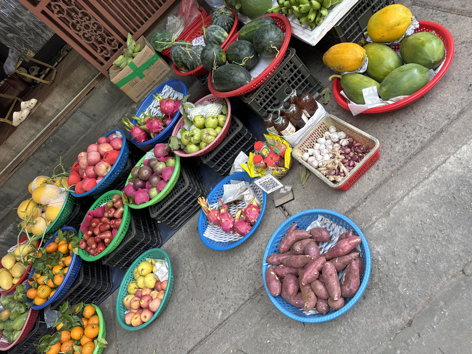 The image depicts a street-side fruit and vegetable market in Đà Nẵng, Vietnam. Several blue, red, and green plastic baskets contain a variety of fruits including mangoes, oranges, red and green apples, rose apples, dragon fruit, guava, watermelons, papayas, and sweet potatoes. Other items are present, including bananas in a box, bottles of liquid (possibly juice or sauce), and a basket containing garlic, shallots, and ginger. The scene is on a concrete sidewalk next to a dark metal gate, likely in a residential or commercial area. There are no people fully visible in the image, although there are shoes present in the upper left.