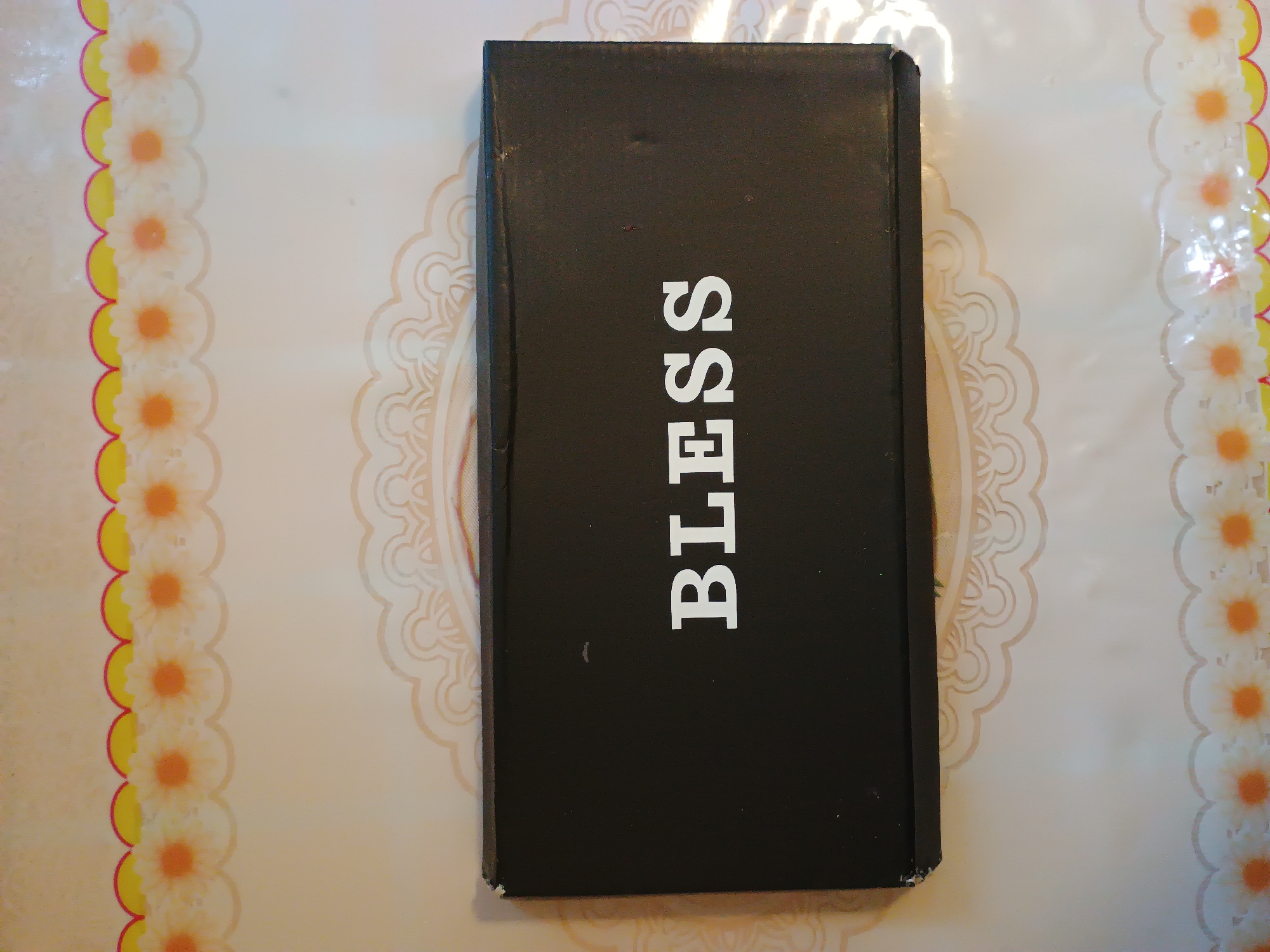 A black box with the word "BLESS" in white capital letters is centered on a decorative tablecloth. The tablecloth has a light-colored background with a repeating pattern of light brown, scalloped lace-like designs. Along the top and bottom edges of the image, there is a border with alternating orange and yellow petal-like shapes interspersed with white daisy-like flowers. The scene is an indoor setting. There are no people or animals present in the image. The image does not contain information that would allow for determining its location in Zaporizhzhia, Ukraine.