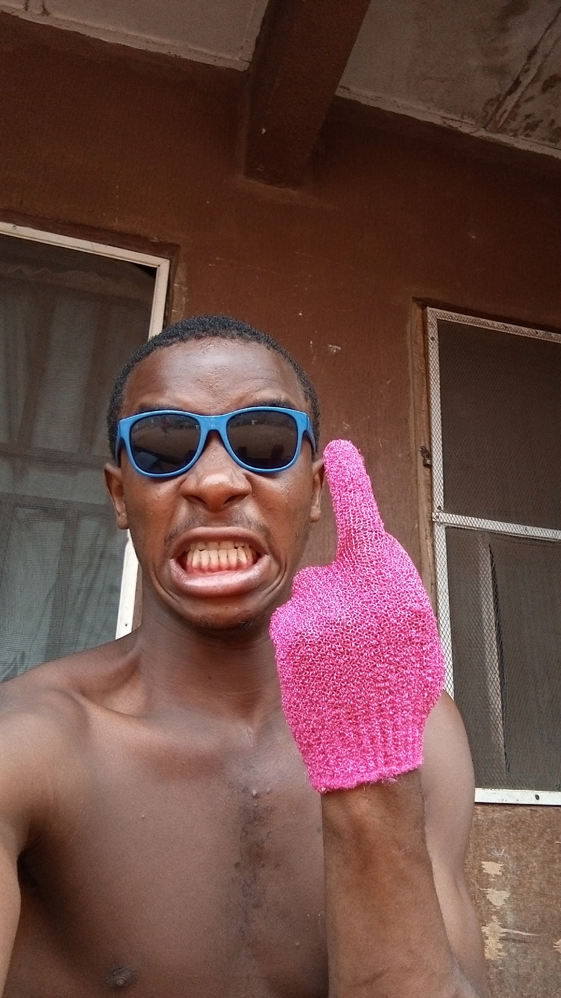 A young Black man is captured in a close-up, comedic portrait, shirtless and wearing bright blue sunglasses. He makes an exaggerated, humorous facial expression, pulling his lips back to reveal his teeth in a wide grimace. His left hand, adorned with a vibrant pink, textured glove (reminiscent of a loofah or exfoliating glove), is raised with the index finger pointing upwards.
The setting appears to be outdoors or in a semi-enclosed space in Onitsha, Nigeria, with a plain, earthy brown wall forming the background. Parts of simple window frames, fitted with mesh screens, are visible on either side of the man, suggesting a practical building structure. The lighting is bright and even, indicative of daytime. The overall mood conveyed by the man's pose and expression is playful and lighthearted. No text is visible in the image.