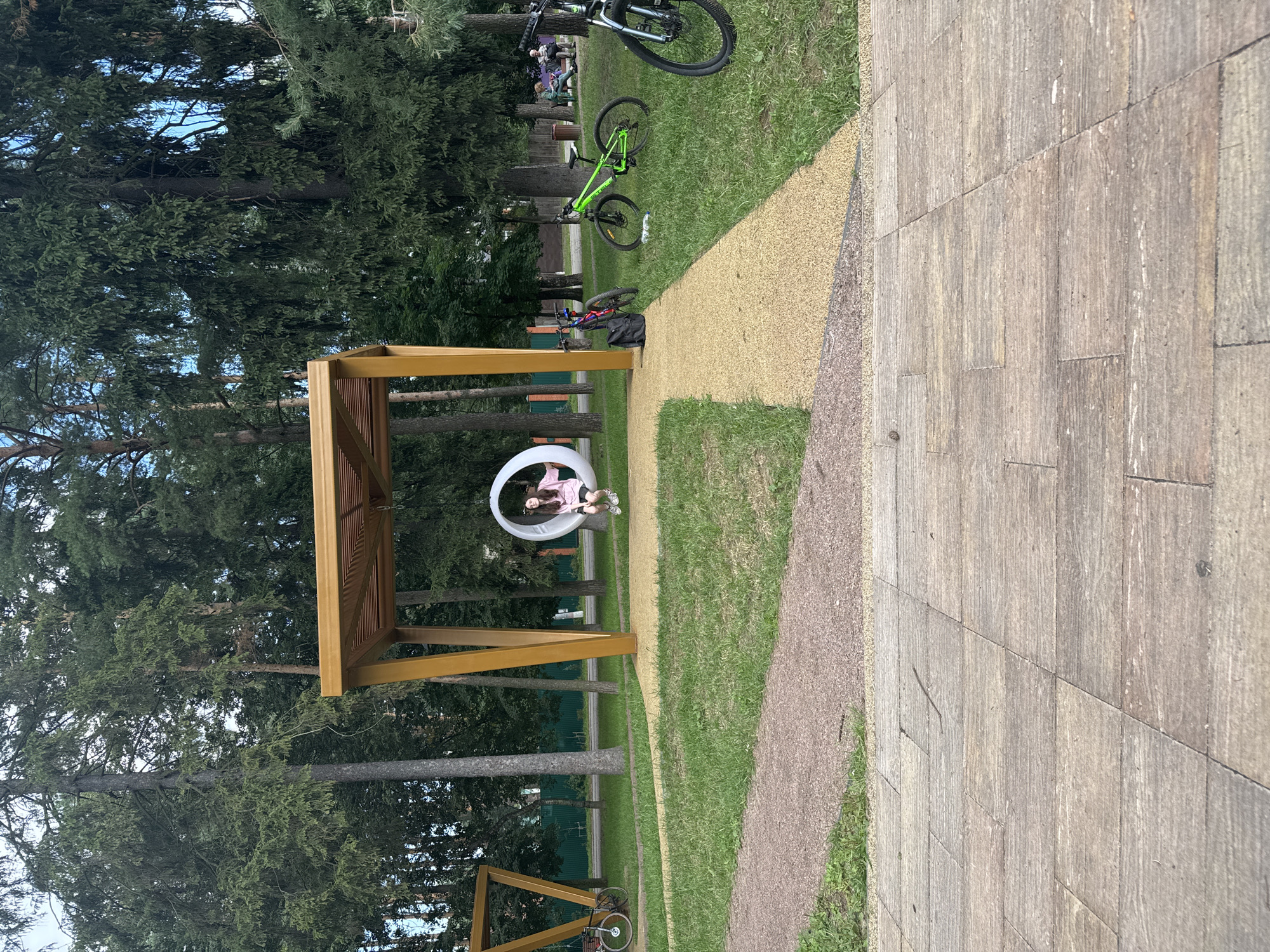 In Il'inskiy, Russia, a scene unfolds in a park-like setting with mature trees framing a grassy area. In the foreground, a paved walkway made of large, rectangular stone slabs stretches across the bottom of the frame. Beyond the walkway, a path of tan-colored gravel curves through the grass. Center-left, a triangular wooden structure with a metal support stands, with a bicycle leaning against its base. Nearby, a young girl with long dark hair, wearing a pink hooded jacket and black shorts, sits on a white, circular swing suspended from a wooden canopy structure. To the right of the canopy, two bicycles are parked on the grass; one is bright green, and the other is darker. Further in the background, a few adults are seated on a bench under the trees. A dark fence runs along the perimeter of the park, with some buildings visible beyond it.