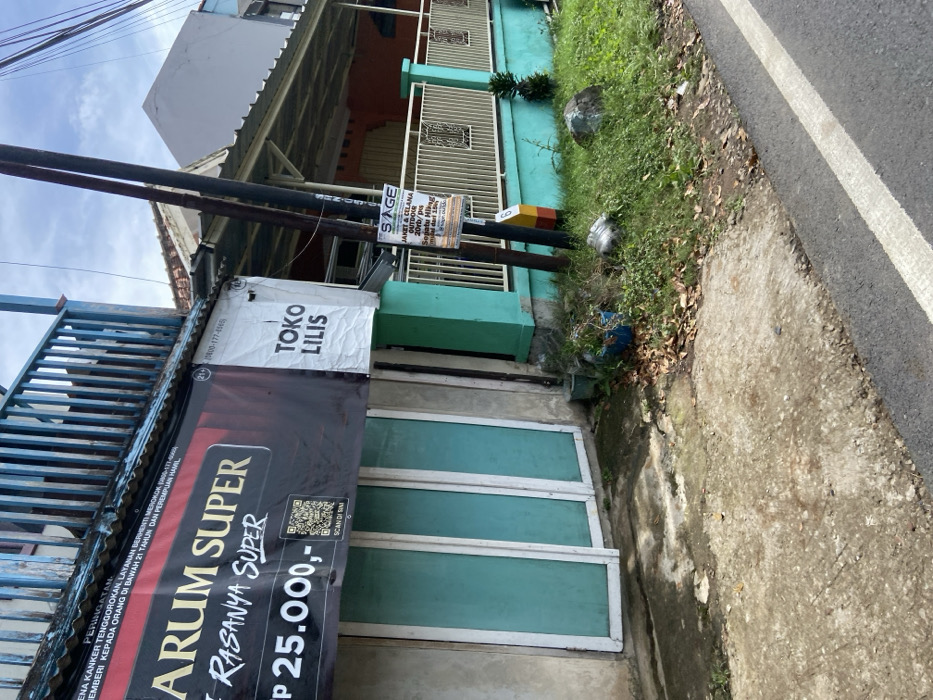 The image shows a street scene in Rancabali, Indonesia. It is a storefront with a blue and white sign that reads "ARUM SUPER / RASANYA SUPER / Rp 25.000,-". A white sign next to the storefront reads "TOKO LILIS". To the right of the store, there is a light pole with a sign advertising "JAKET & CELANA OUTDOOR 20% DIS" with the logo "SAGE". The storefront has a set of four glass doors with a white frame. The storefront is next to a concrete sidewalk with a yellow painted curb separating the sidewalk from a paved road. There is grass and some dead leaves on the sidewalk and a small potted plant in the foreground. The image was taken on a clear day. The image is slightly blurry and the colors are muted. The location in the city of Rancabali, Indonesia, is based on coordinates (latitude: -7.12, longitude: 107.432).