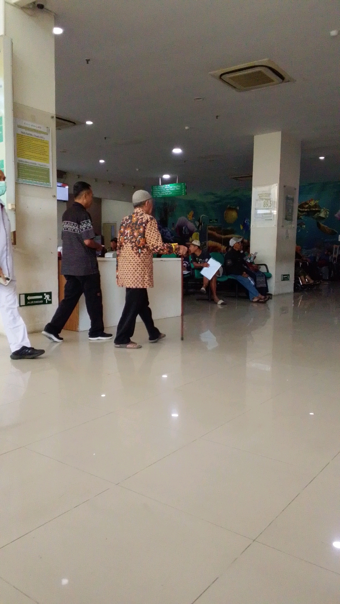 The image depicts the interior of a brightly lit, modern building, possibly a hospital or clinic, in Pabelan, Indonesia. The floor is tiled with large, pale beige squares, reflecting the overhead lighting and the interior decor.
In the foreground, two men are walking side-by-side. The man on the left wears a dark, patterned short-sleeved shirt and black pants. The man on the right, an elderly individual, wears a patterned long-sleeved shirt, black pants, and a light-colored cap. He is using a walking cane. To the far left, a person in white clothing, possibly a medical professional, is partially visible.
Further into the scene, several other individuals are seated, appearing to be waiting. One person is holding a document. The background features a counter, a television screen displaying information, and a wall decorated with a vibrant mural of underwater scenes, complete with fish and turtles. Signs with green text are mounted on the wall, indicating directions or information. An air conditioning unit is visible in the ceiling. The overall atmosphere is one of a public waiting area, with people engaged in their own activities or observing their surroundings. The lighting suggests it is daytime.