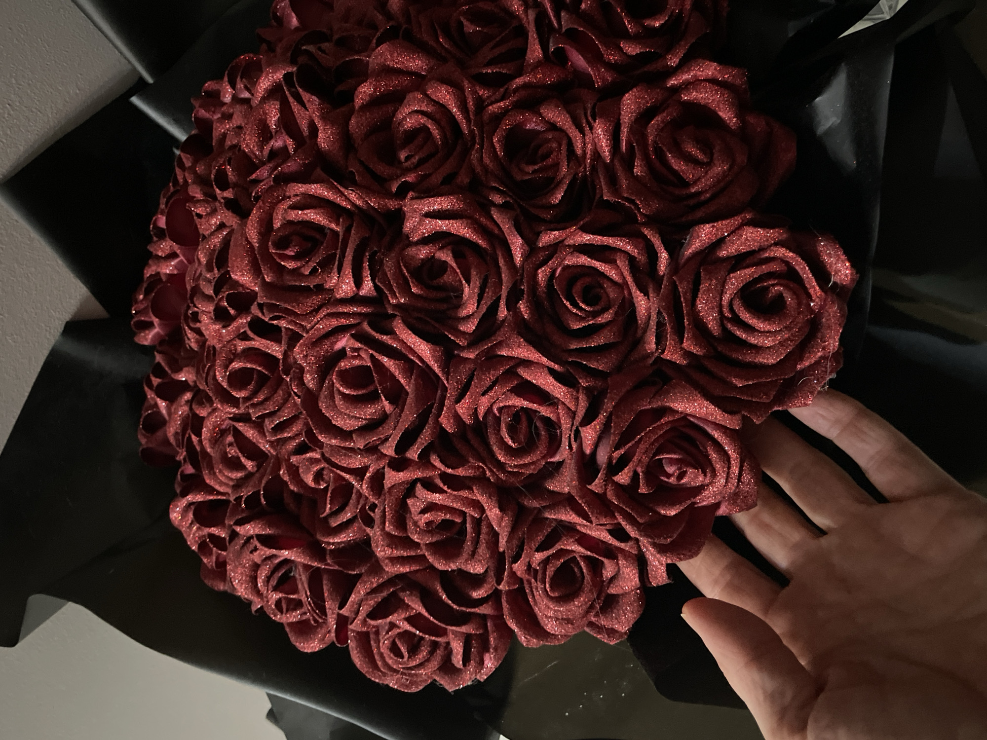 A bouquet of red glitter roses is presented, with a human hand partially visible in the foreground. The roses are densely packed and appear to be made of fabric. They are arranged in a round bouquet and are wrapped in black material. The background is indistinct, with a wall visible in the upper left corner. There is no indication of the location being in Sochi, Russia.
