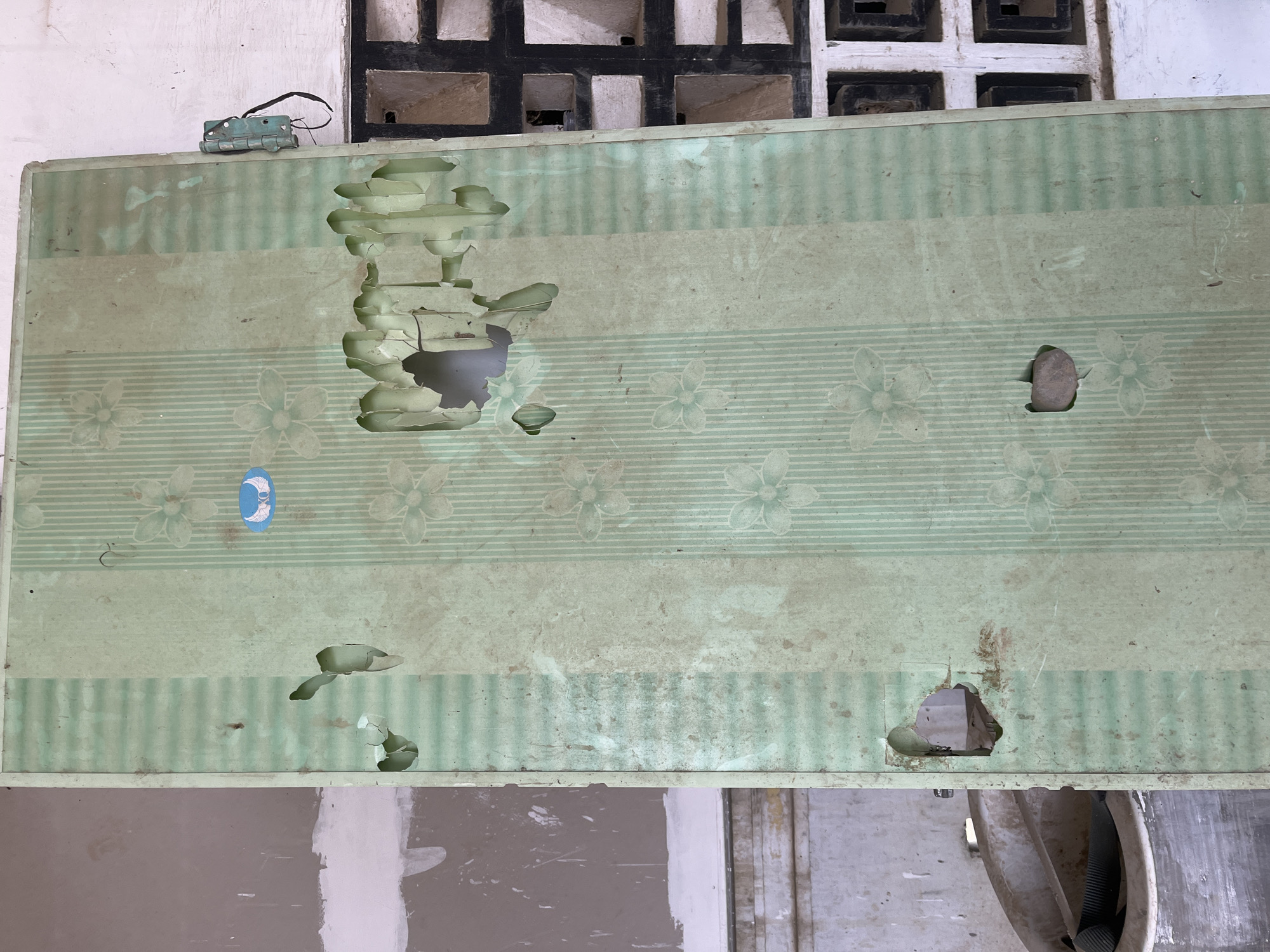 The image shows a close-up, slightly overhead view of a damaged, light green ceiling panel with a striped and floral pattern. The panel is dirty and has several large holes where the material has broken away, revealing a darker interior. One hole is particularly jagged and large, located in the upper left quadrant of the panel. Another smaller, rounded hole is in the center right, with a small, greyish object, possibly a pebble, lodged in it. A blue and white circular sticker depicting a crescent moon and stars is visible in the upper left portion of the panel.
Above the ceiling panel, a white wall is visible, and at the top of the image, a section of a black and white patterned ventilation brick structure can be seen. A small, metallic, blue object, possibly a hinge or part of a latch, with black wires attached, is resting on the wall just above the ceiling panel.
The lower portion of the image shows a blurry view of walls and some piping or ductwork. The overall impression is one of disrepair and neglect in an indoor setting. The lighting appears to be neutral, suggesting it is daytime, but there are no clear indicators of weather conditions. The scene seems to be located in an interior space, possibly a room or a building undergoing renovation or demolition. No people are visible in the image.