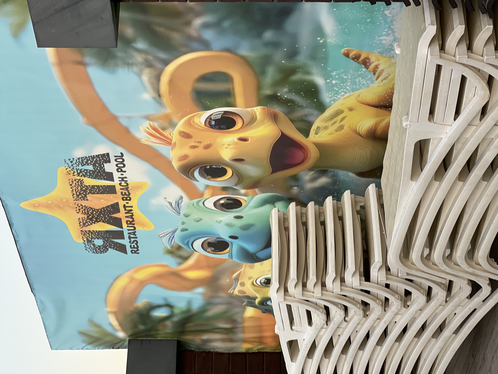 In Fontanka, Ukraine, a close-up, eye-level shot captures a large banner depicting cartoon dinosaurs and a logo that reads "ЯХТА RESTAURANT BEACH POOL" under a yellow starfish. In the foreground, a stack of white plastic lounge chairs is neatly arranged, partially obscuring the banner. The banner displays a bright blue sky and a yellow water slide. The banner is a promotional advertisement for the restaurant, beach, and pool. The scene suggests a summer vacation or resort setting.
