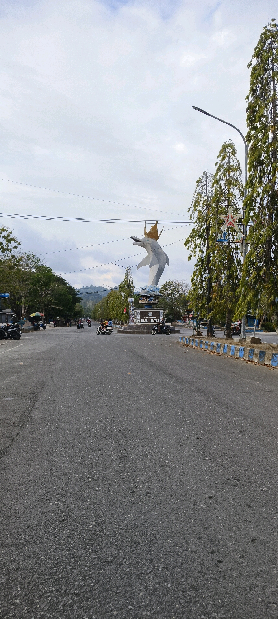 A wide, paved road in Waisai, Indonesia, leads towards a prominent roundabout featuring a large dolphin monument. The dolphin, depicted leaping with a crown on its head, is a gray and white sculpture. Behind the monument, a sign reads "KABUPATEN RAJA AMPAT."
The road is asphalt and shows some wear and tear. Traffic is light, with several motorcycles and a few cars visible. On the left side of the road, there are trees and buildings, including a roadside stall with colorful umbrellas. On the right, a row of tall, slender trees lines the median of the roundabout. A street lamp is visible above the trees.
The sky is overcast with soft, diffused light. The overall impression is of a quiet day in a coastal town, with the dolphin monument serving as a central landmark. The image is captured from a low angle, emphasizing the road and the monument ahead.