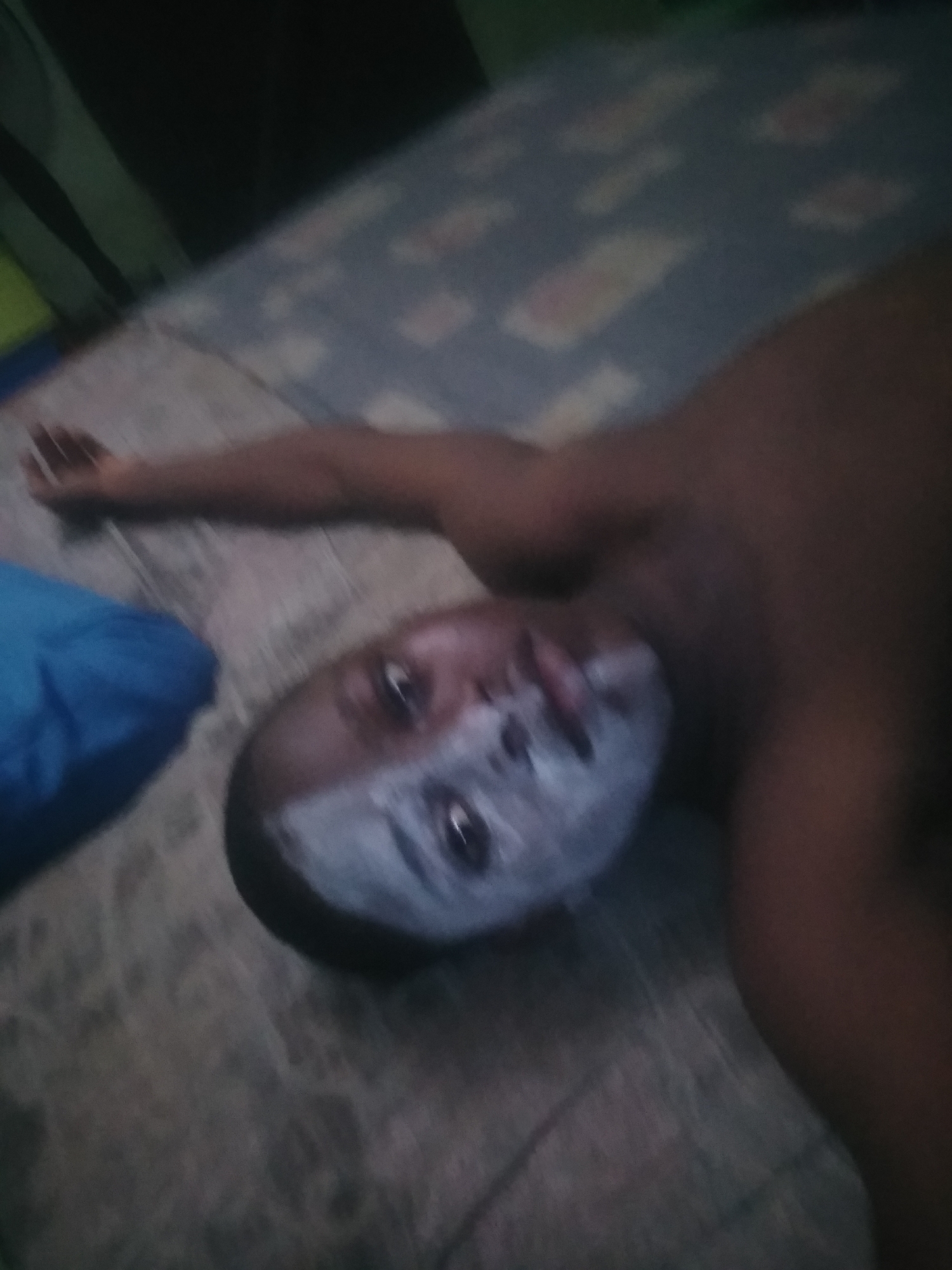 A dark-skinned person is depicted lying on their back, viewed from a slightly elevated and tilted perspective. Their face is partially covered with a white substance, possibly a face mask, makeup, or paint, extending from their nose across their cheeks and chin. Their eyes are open and look directly towards the camera. The person appears to be shirtless, with their left arm extended upwards. A crumpled blue pillow is visible near their left shoulder. They are lying on a bed or mattress with a faded, patterned sheet in shades of blue and gray. The background beyond the bed is dark and blurry, suggesting a dimly lit indoor environment, possibly a bedroom. The time of day appears to be evening or night due to the low light. No specific location cues or visible text are present. The person's expression is neutral, and the white facial covering adds a distinct, somewhat ambiguous detail to the scene.