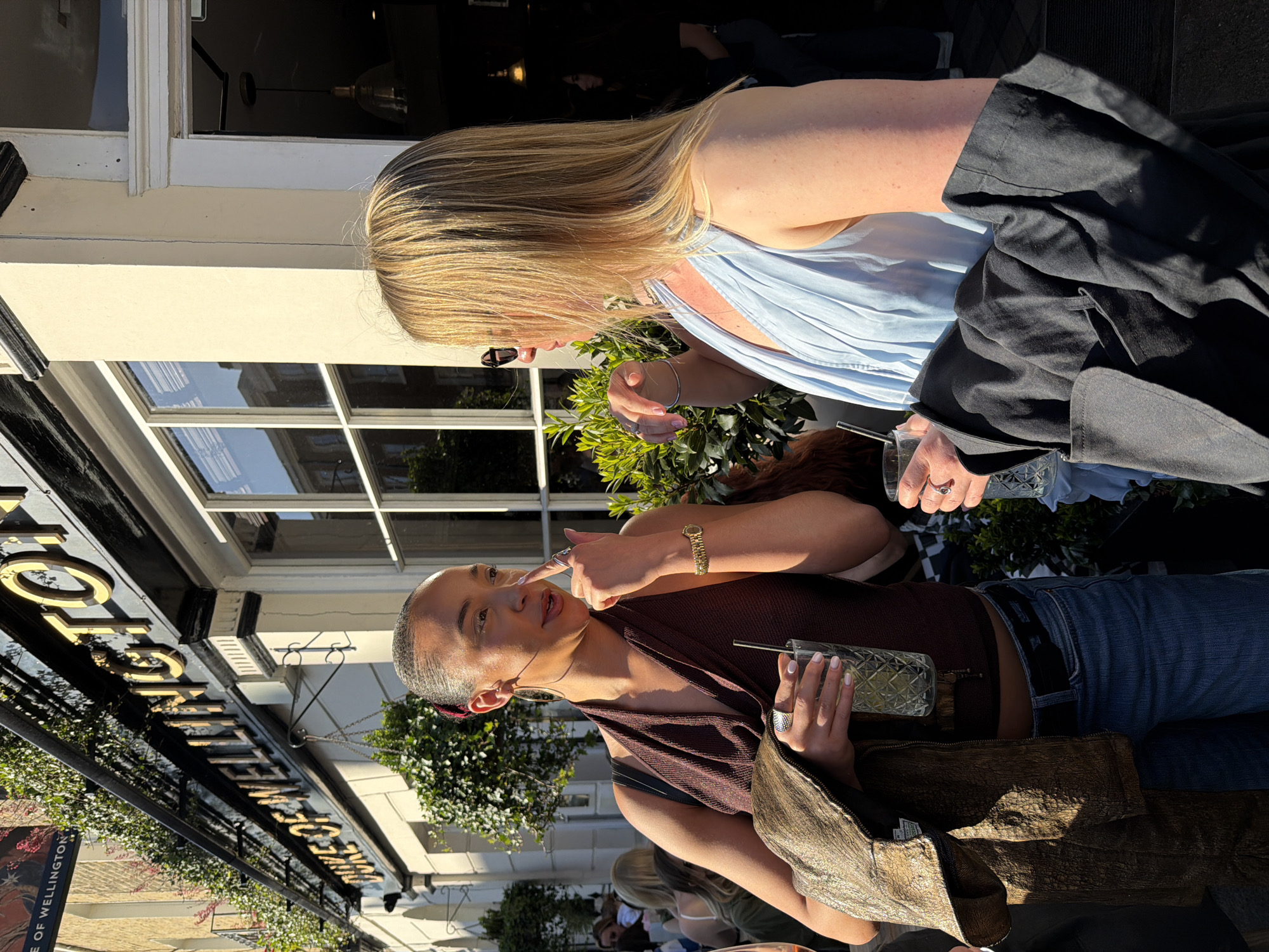 The image captures two individuals, a blonde-haired woman and a darker-haired person, sitting at a table outdoors. The woman with blonde hair is wearing a light blue top and a black jacket, while the other person is wearing a brown halter top and jeans. Both appear to be holding drinks. They seem to be engaged in a conversation.
The backdrop reveals that they are located near the 'Duke of Wellington' pub, which is indicated by the signage. This suggests the setting is in London, United Kingdom. There are plants decorating the exterior of the building and window reflections suggesting other buildings across the street. Other patrons can be seen in the background.