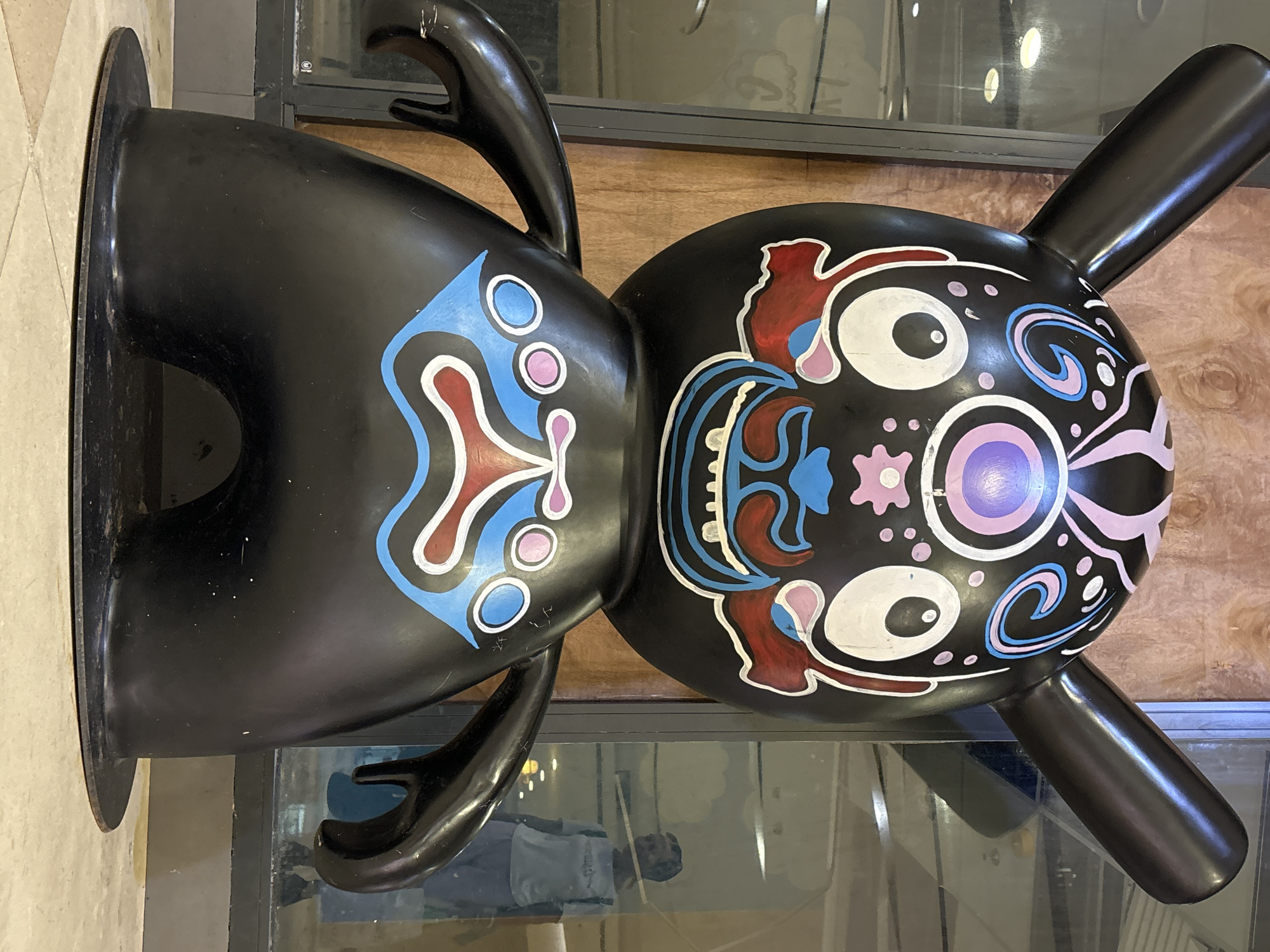 A full shot captures a large, black, stylized cartoon figure with prominent ears and colorful, abstract facial markings, standing on a circular base. The figure's face features large white eyes with purple pupils, a red and blue patterned mouth, and pink and blue swirl designs on its forehead. Its body is adorned with blue and pink shapes. The figure is positioned in front of a wooden-paneled wall and a large glass window, through which a blurred reflection of a person wearing a light-colored shirt and dark pants is visible. The scene is lit with overhead lights, and the floor is tiled with a beige and cream pattern. The overall impression is that of an art installation, possibly in a public space like a mall or exhibition area in Tianya District, China.