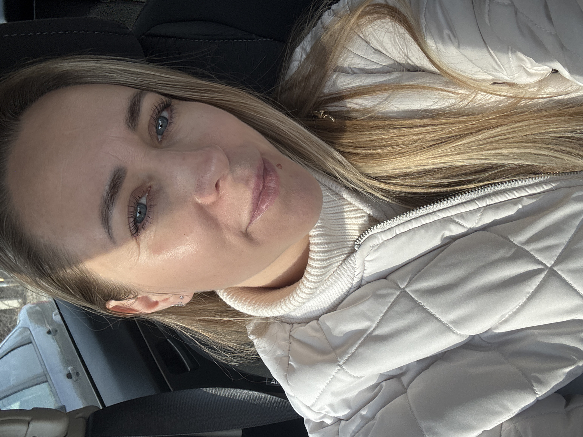 A woman with blonde hair and blue eyes is taking a selfie indoors, likely in a car. She is wearing a white quilted jacket and a white turtleneck sweater. The background shows elements of a vehicle interior. There is no indication of the location being Chaikovsky, Russia, or any other specific city.