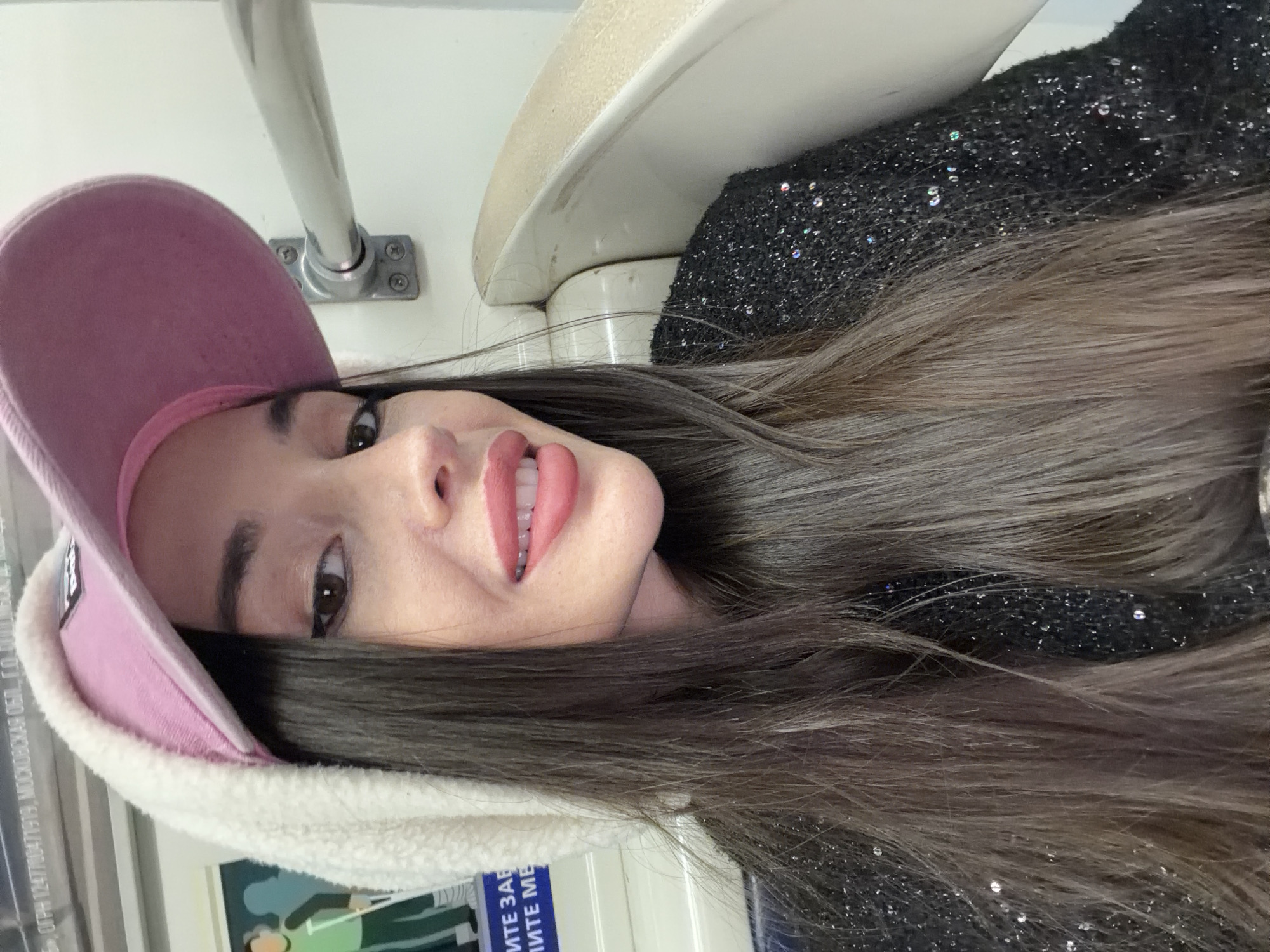 A woman is looking at the camera. She wears a pink baseball cap with a white, furry edge. She has long brown hair. The woman wears a black shirt with glitter. She is on public transportation, possibly in a subway car, judging by the pole and white seat above her. Information printed on the wall reads "ОГРН 1247700547919, МОСКОВСКАЯ ОБЛ., ГОРОД..." The location is identified by the Russian language text and the interior of the transportation, which is most likely in Saint Petersburg, Russia.